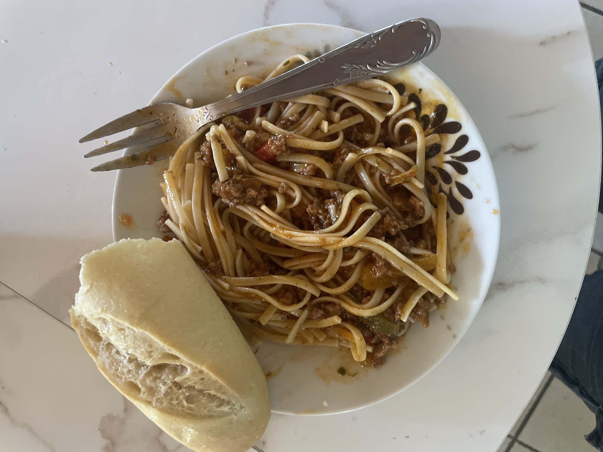 The image shows a plate of pasta with meat sauce and a piece of bread. A fork rests on top of the pasta. The plate is white with a decorative brown pattern around the rim. The pasta appears to be linguine or spaghetti, and the sauce contains ground meat and possibly some chopped vegetables. The bread is a crusty baguette that has been sliced open. The setting is a white table with a marble-like pattern. The lighting suggests it is daytime, but no specific time can be determined. There are no people visible in the image. The overall impression is of a meal being served.