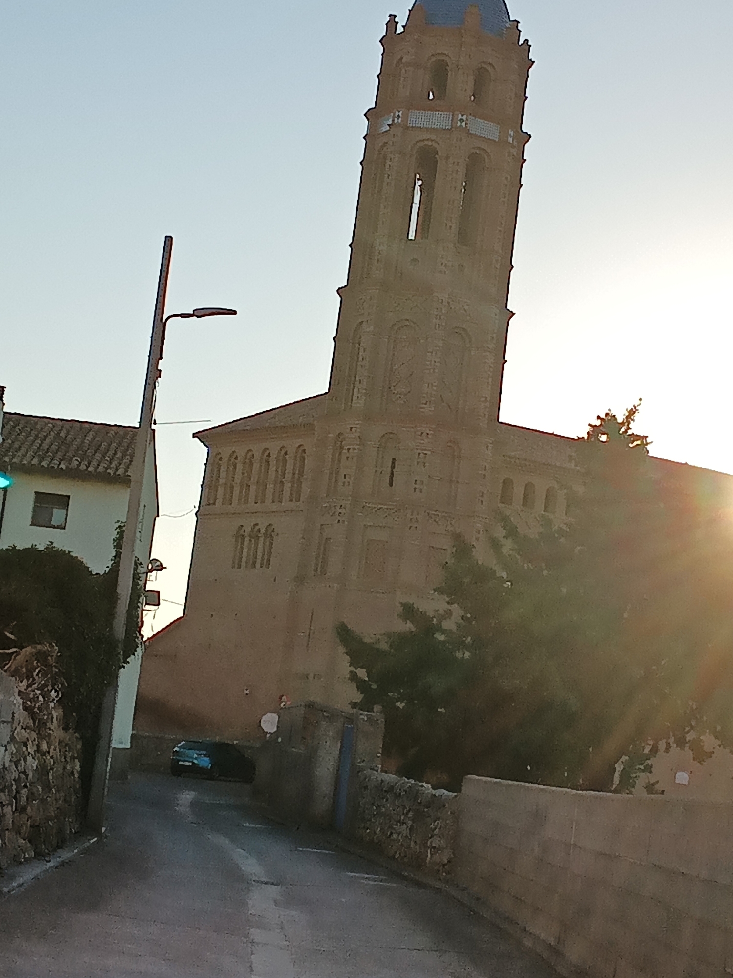 The image shows a street scene in the city of Mainar, Spain. A tall brick church tower with a blue-tiled roof dominates the background. The tower has several arched openings and decorative brickwork. To the left of the tower, a white building with a tiled roof and a small window is visible. A street lamp with a modern fixture stands in front of this building. In the foreground, a narrow, paved street curves to the left, flanked by stone walls and vegetation. A dark-colored car is parked on the street. The sun is setting, casting a warm glow and lens flare across the scene. The sky is clear and pale blue.