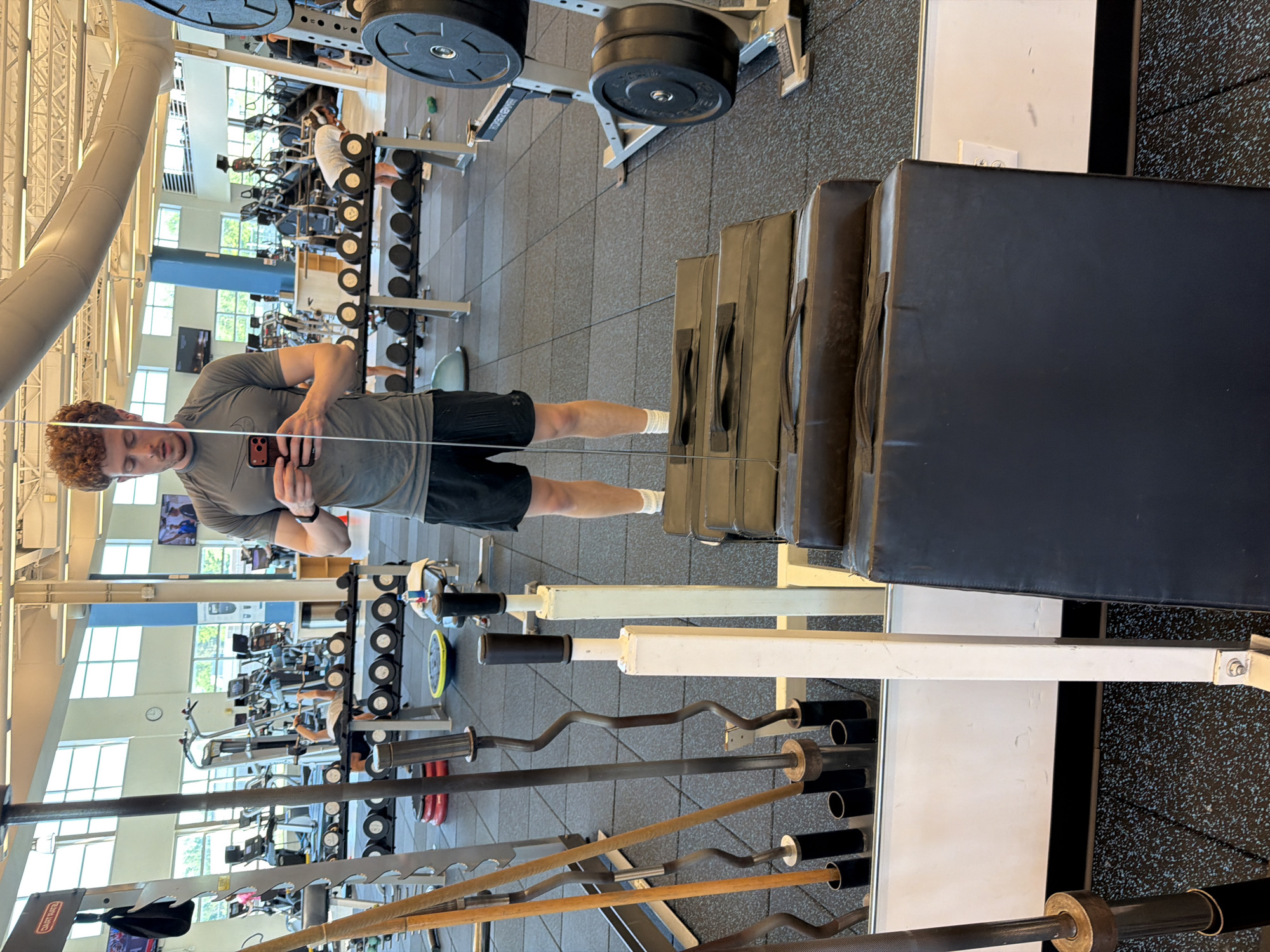 A young man with curly, reddish-brown hair and a light complexion is standing in a gym, taking a selfie in a mirror. He is wearing a grey, short-sleeved t-shirt with a Nike logo on the left chest and black athletic shorts. He has on white socks and dark athletic shoes. The man is holding a smartphone in his right hand, with his left hand near his waist.
The gym environment is visible in the reflection and background. Racks of weights, barbells, and weight plates are present. In the foreground, stacked on the floor, are several large, black padded mats. To the left of the man, in the reflection, are several barbells stored in a rack. In the background, other gym equipment like treadmills and elliptical machines can be seen, along with other gym-goers. The flooring is a dark, textured rubber material. The overall lighting suggests it is daytime, with natural light coming from large windows in the background. The gym appears to be well-equipped and spacious. This image was taken in Beachwood, United States of America.