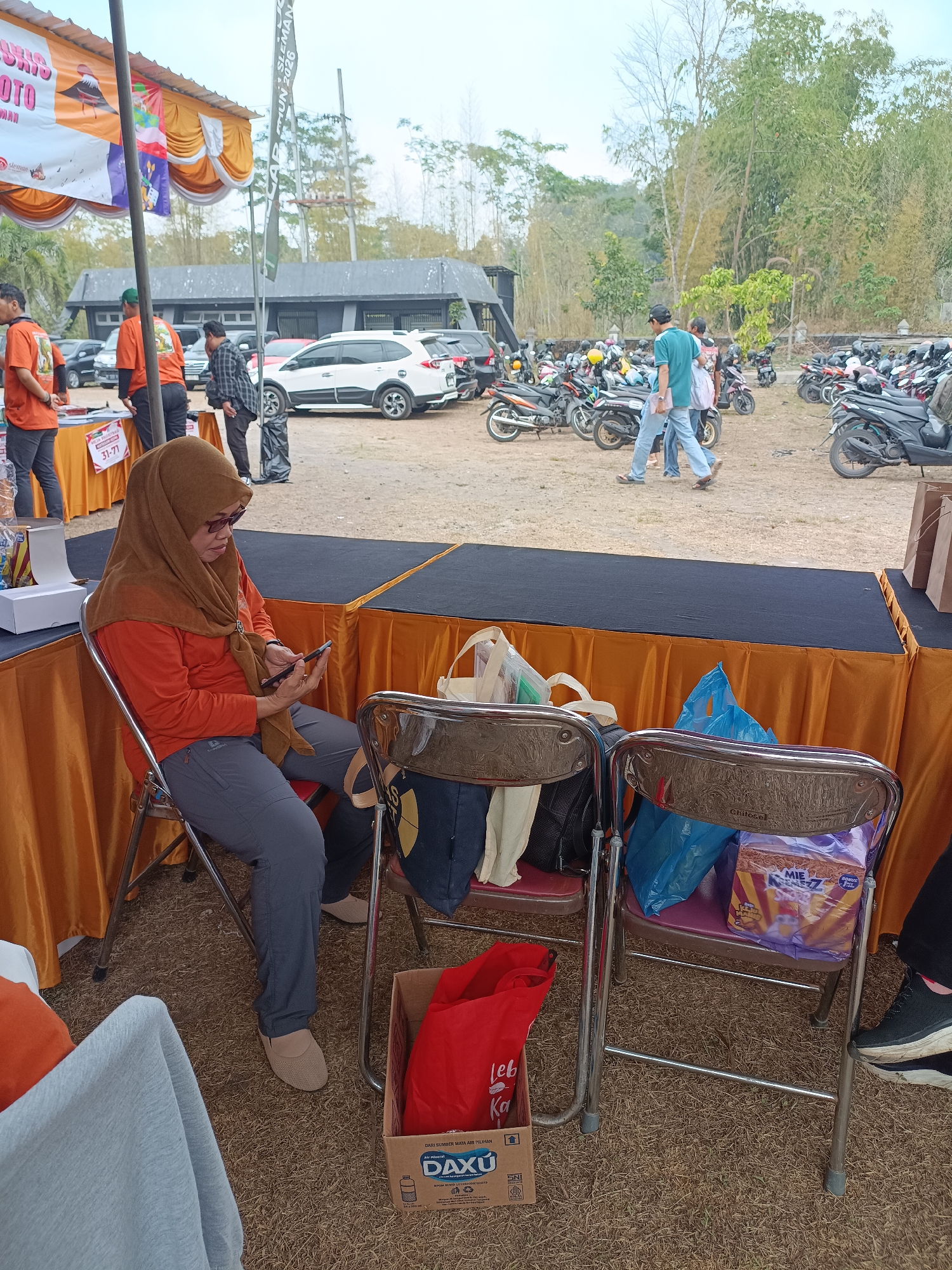 A woman wearing a brown hijab and an orange shirt sits on a metal chair at an outdoor event in Hargobinangun, Indonesia. She is looking at her phone and holding it in her hands. She wears grey pants and light brown shoes. The chair is next to a table covered with an orange cloth. On the table, there are two plastic bags and a box. Behind the woman, there are other chairs and people, as well as many parked motorcycles. The event appears to be taking place in a grassy area with trees in the background. The sky is overcast.