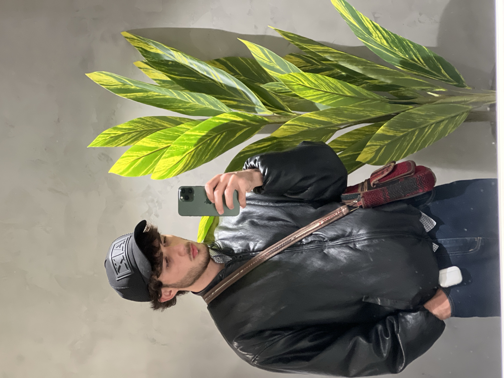 A young man wearing a black leather jacket, a black baseball cap with a logo, and jeans is taking a selfie with his smartphone. He has a cross-body bag with a patterned red and black fabric. He is standing in front of a plant with long, green and yellow striped leaves. The background appears to be a grey wall. The location is likely indoors in Yerevan, Armenia, as per the user's request for the city and country to be identified.