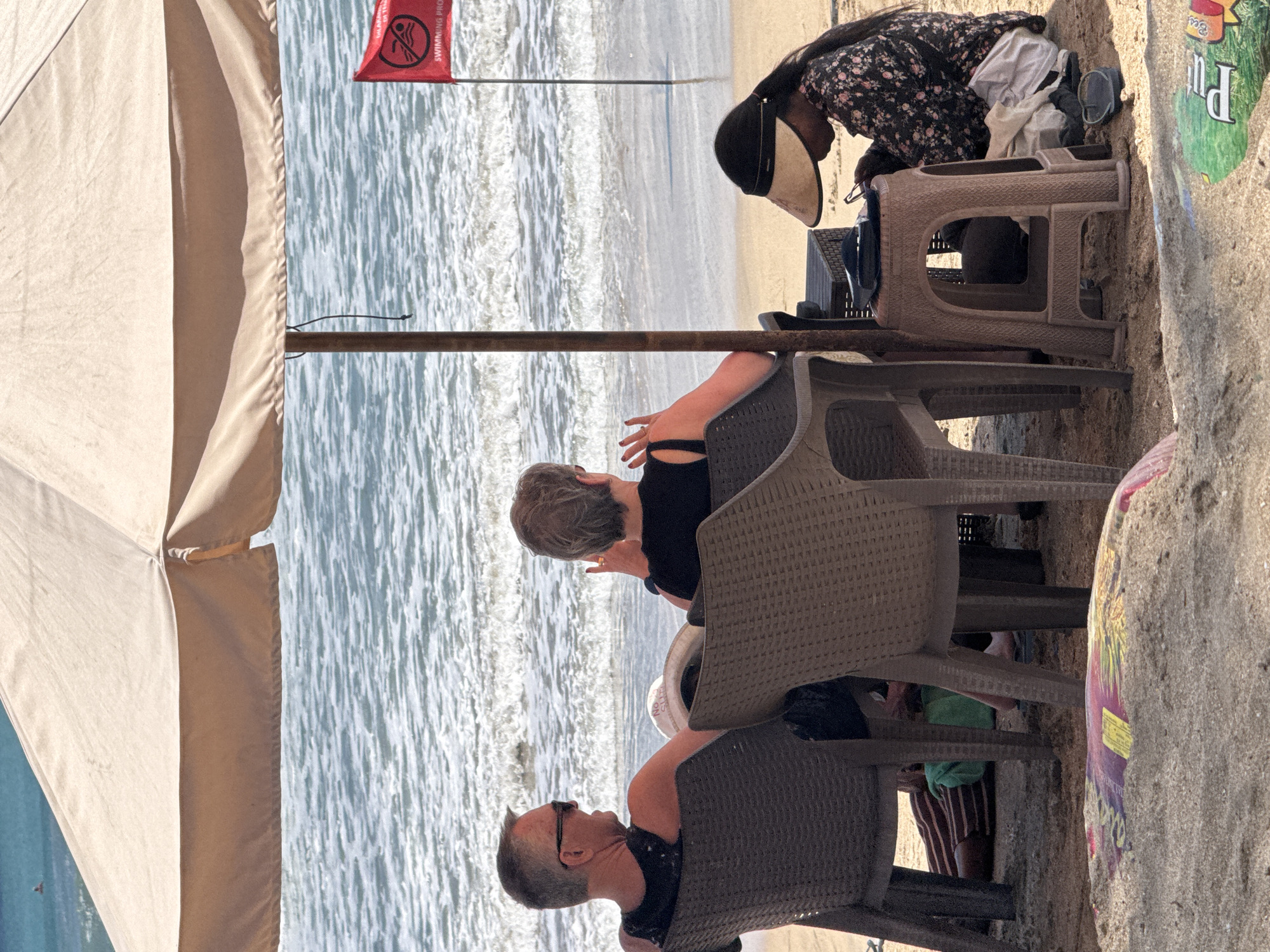 The image shows a beach scene in Kuta, Indonesia, likely under a large beach umbrella. In the foreground, a person in a floral-patterned shirt and a sun visor sits on the sand, appearing to be engaged with a small electronic device and holding sunglasses. Beside them, two plastic chairs are occupied by a man with sunglasses and a woman with short hair, both facing the ocean. The man is wearing a black tank top, and the woman is wearing a black top. The ocean is visible in the background with gentle waves and a red flag indicating that swimming is prohibited. The sand is a light brown color, and there are some discarded items, including what appears to be a portion of a surf mat or board, in the lower foreground. The overall atmosphere is relaxed, with people enjoying the beach under the shade.