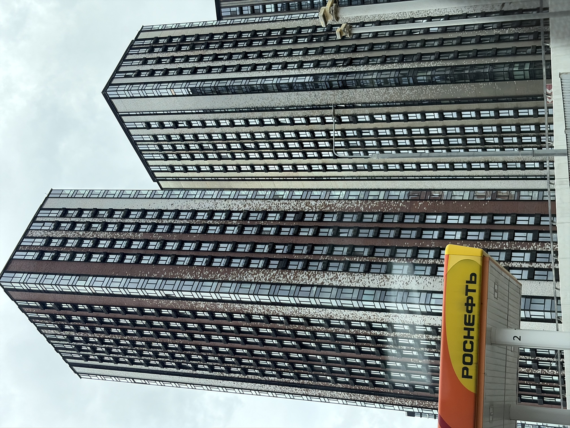 The image shows a view of two modern, tall residential buildings in Moscow, Russia, under an overcast sky. In the foreground, a section of a gas station canopy is visible on the left side of the frame. The canopy is orange, yellow, and red, with the Russian text "РОСНЕФТЬ" (Rosneft) printed in black. A white pillar with the number "2" in black is partially visible beneath the canopy. The buildings feature a facade of glass windows and brickwork in various shades of brown and beige. The windows are arranged in a grid pattern, with some appearing to be balconies. The lighting suggests it is daytime, but the cloudy conditions create a diffused light. No people are visible in the image.