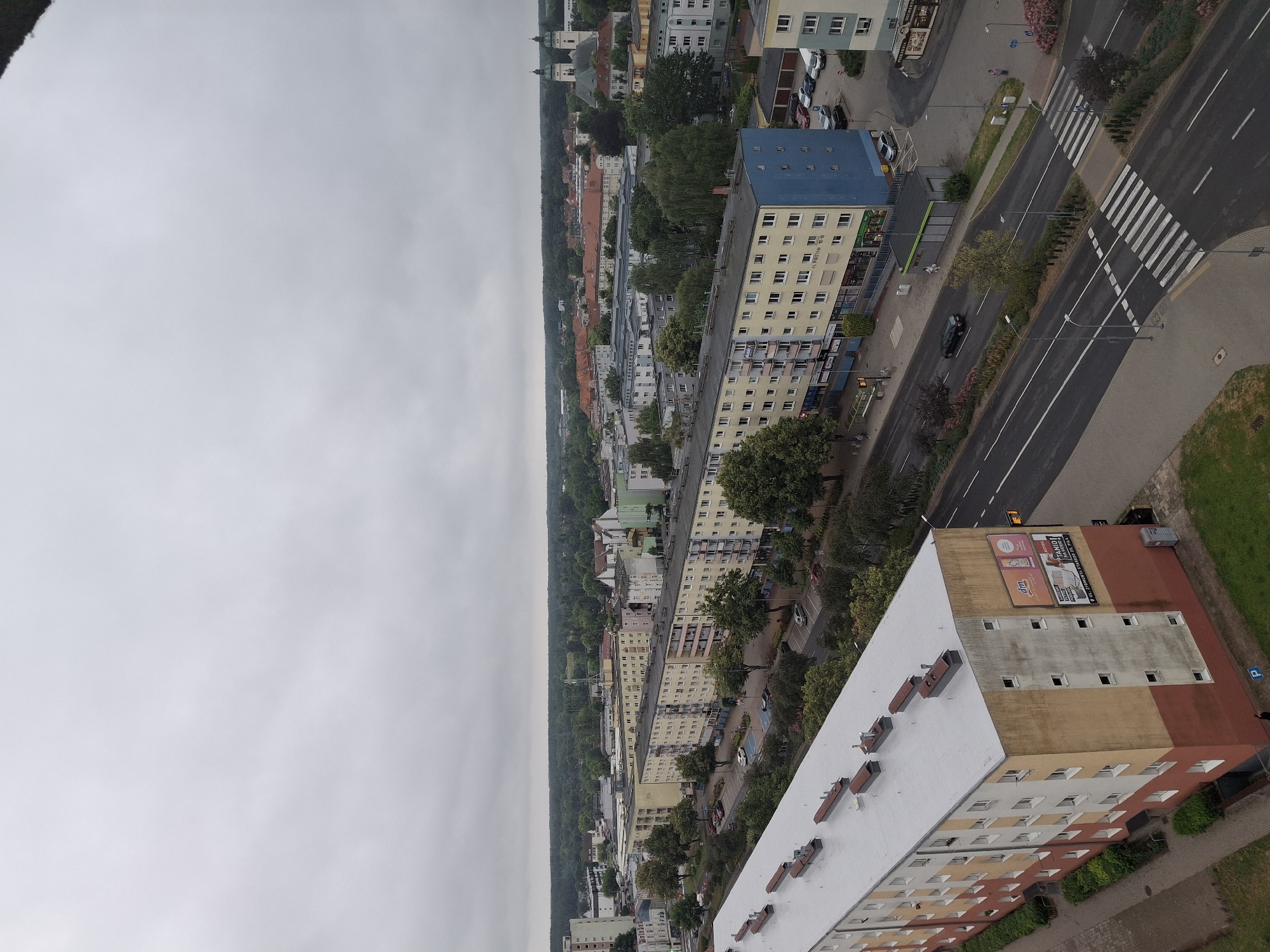 The image depicts an overcast day in the city of Piła, Poland, showing a view of a residential area and surrounding buildings. In the foreground, a long, multi-story apartment building with a light-colored roof runs diagonally across the frame. The building has a facade of yellow and red sections with numerous windows. Adjacent to this building is a street with cars, parked vehicles, a pedestrian crossing, and trees lining the side.
Further into the distance, several other apartment buildings are visible, some with yellow facades and others with a mix of colors including green and blue. Beyond the residential blocks, a denser area of buildings suggests a more commercial or older part of the city. Distinctive structures include a church with twin steeples and a large, red-roofed building. The background is filled with a dense line of trees, indicating a forest or large park bordering the city. The sky is uniformly grey and cloudy, typical of an overcast day. A few people are visible on the sidewalks and streets, walking or standing. The overall scene is one of urban residential development with natural elements present in the background.