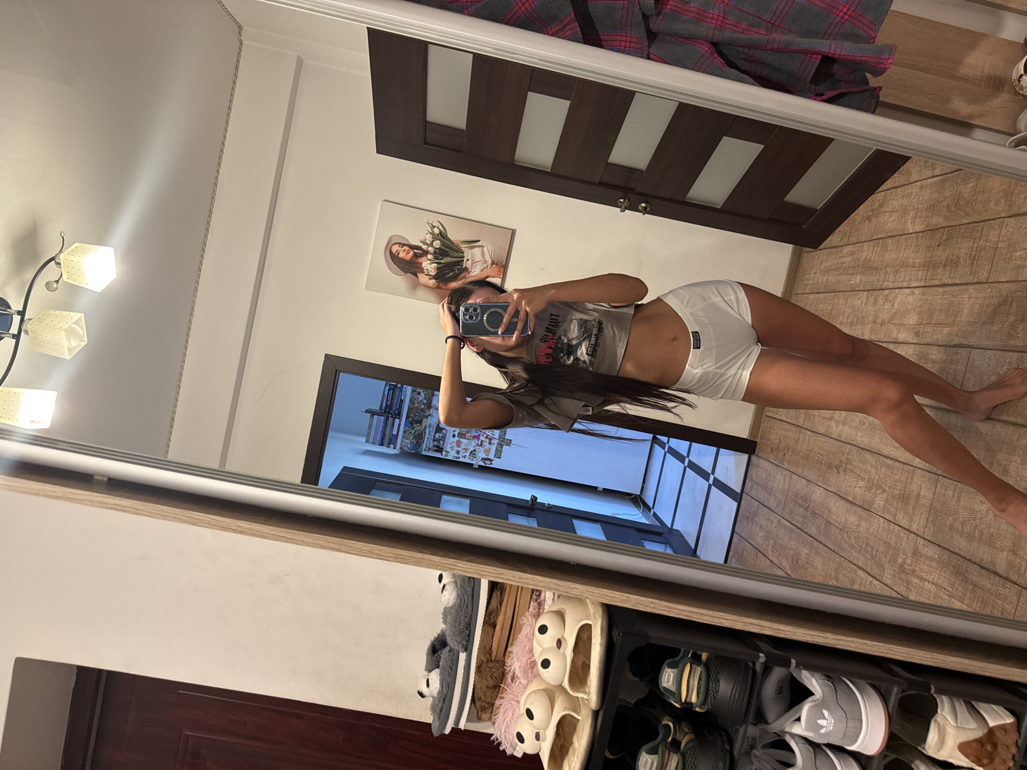 In a full-length mirror reflection, a young woman stands in what appears to be a bedroom or dressing area. She is wearing a cropped t-shirt and white shorts, and is holding a smartphone to take the photo. The room has wooden flooring and a dark brown door with vertical white panels. On the wall, a framed picture depicts a woman with a hat and flowers. A shoe rack is visible at the bottom of the mirror, filled with various pairs of shoes and slippers, including novelty slippers with large googly eyes. A large plaid blanket is draped over a surface in the upper portion of the reflection. The lighting suggests it is daytime, possibly indoors with artificial light sources contributing to the overall illumination. The image is taken from a low angle, looking up towards the woman. There is no discernible text in the image, other than a small label on the woman's shorts which is too blurry to read.