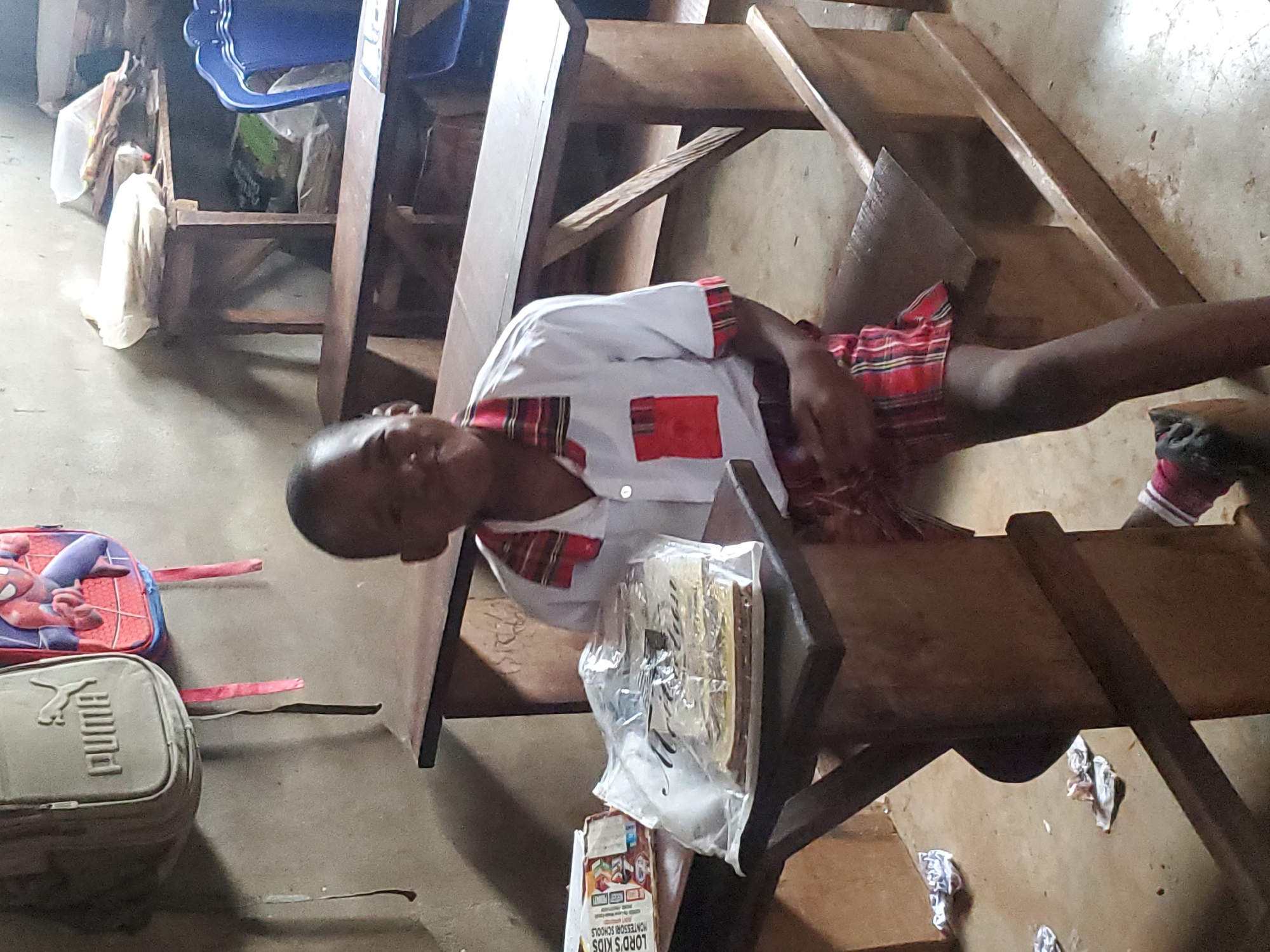 A boy wearing a white shirt with red accents and a red and black checkered skirt sits at a wooden desk in a room in Ukwu-Abwa, Nigeria. A plastic bag containing papers and a package labeled "LORD'S KIDS MONTESSORI SCHOOLS" sit on the desk. A Puma backpack and a Spiderman backpack rest on the floor nearby. Other school desks and chairs are visible in the background. The floor is concrete.