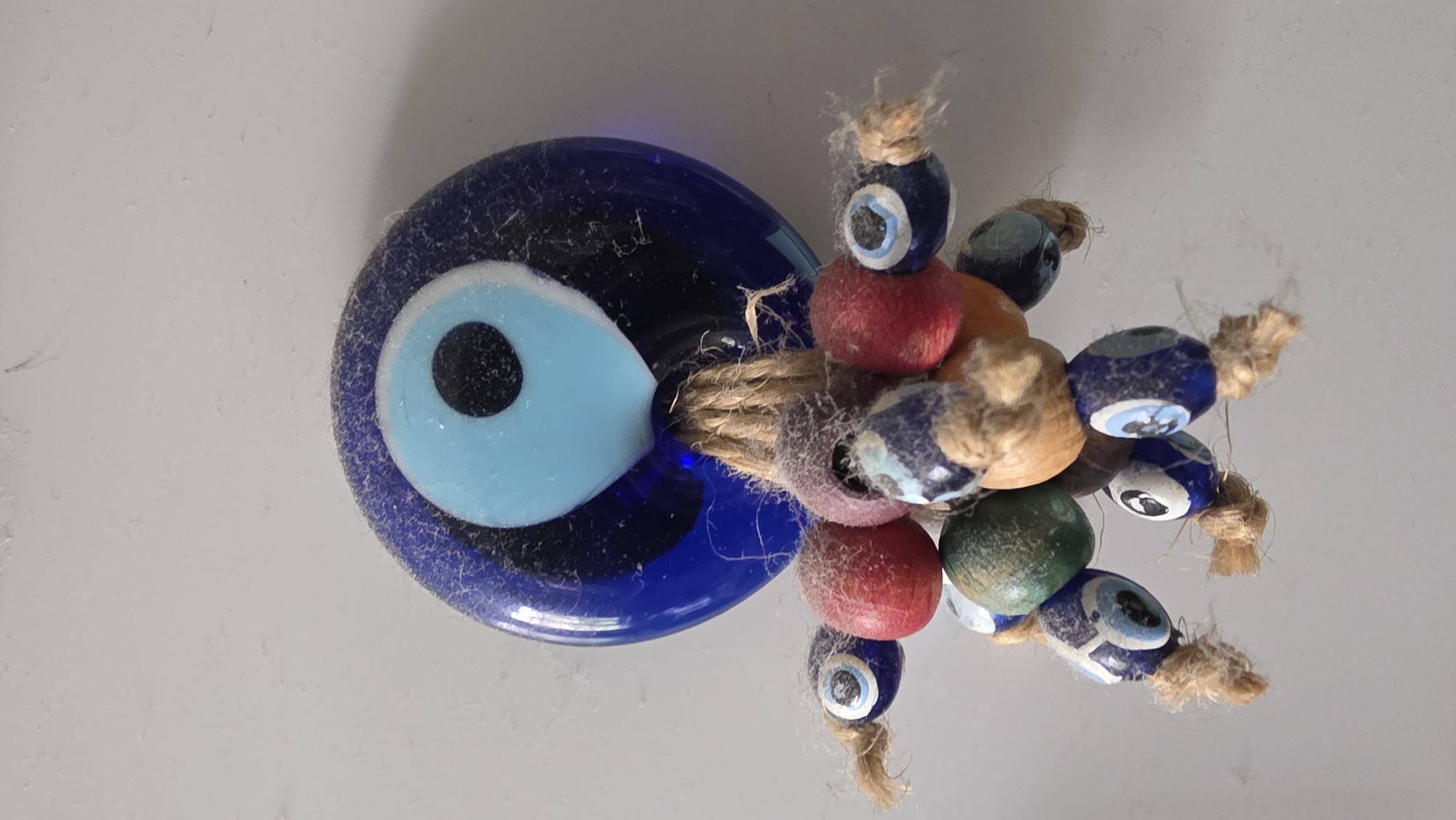 This media file captures a close-up, static shot of a traditional evil eye charm, known as a Nazar Boncuğu, and associated decorative beads. The main subject is a large, glossy blue glass evil eye, featuring concentric circles of dark blue, light blue, and black to form the iris and pupil. This main charm is noticeably dusty or has fine fibers adhering to its surface.
Attached to the larger evil eye, likely by a string or twine, is a cluster of smaller, multi-colored beads. These smaller beads are threaded together with a natural, rustic-looking jute or hemp twine, which also shows some loose fibers. Among these smaller beads, some are also miniature evil eye designs (blue and white/light blue), while others are solid colors including red, green, and natural wood tones.
The object is positioned against a light, possibly white or light grey, flat surface that appears somewhat textured or lightly dusted. The lighting suggests an indoor setting, with no specific time of day or weather conditions discernible. There is no visible activity or event taking place, as it's a still life image. No text is visible in the image.
The presence of the Nazar Boncuğu, common in the Balkans, aligns with the provided location context of Blagoevgrad, Bulgaria. The image is oriented with the main evil eye on the left and the cluster of beads extending to the right.