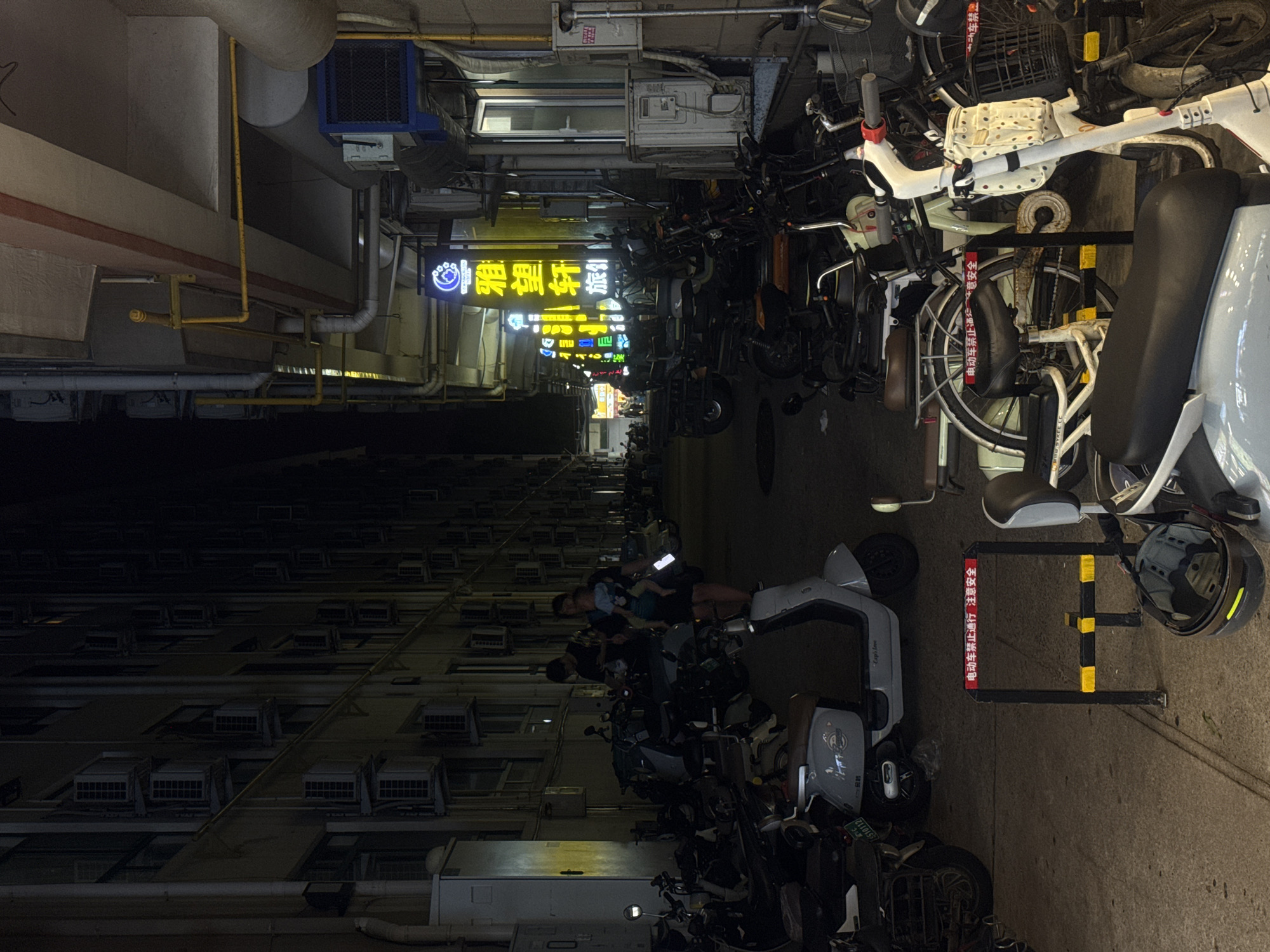The image shows a nighttime street scene in Qinhuangdao, China. The street is lined with multi-story buildings adorned with numerous air conditioning units. A variety of parked electric scooters and motorcycles fill the foreground and midground. Two individuals, an adult male and an adult female holding a baby, are walking in the midground, passing by the parked vehicles. The scene is illuminated by streetlights and neon signs, one of which displays Chinese characters. A red and yellow barrier with Chinese text indicating "Electric vehicles forbidden to pass, safety first" is visible in the foreground. The overall environment appears to be a commercial or residential area within the city.