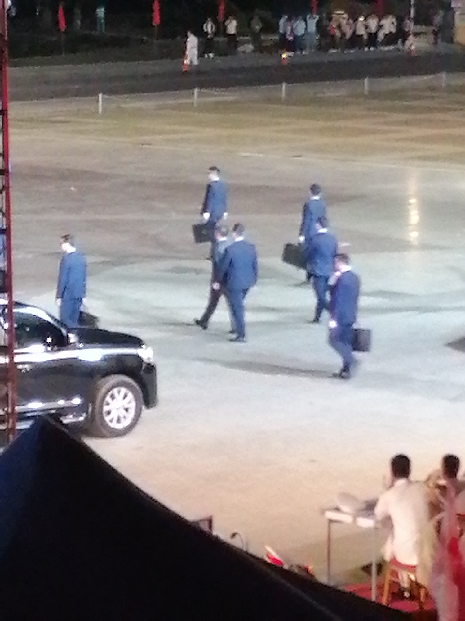 The image shows a group of people, mostly men in suits, walking across an outdoor, paved area. One man in a blue suit stands next to a black SUV. Several other men, also in dark suits, are carrying briefcases and walking away from the viewer. In the background, there are people standing and watching, some holding red flags. A few red flags are also visible on poles in the distance. The lighting suggests it is daytime, possibly late afternoon or early evening, given the shadows. The ground appears to be a large, open space, possibly a plaza or an event ground. The presence of the SUV and the formal attire of the men suggest a formal event or arrival/departure. The overall mood appears to be serious or business-like. No visible text is present in the image. The location context is Da Lat, Vietnam.