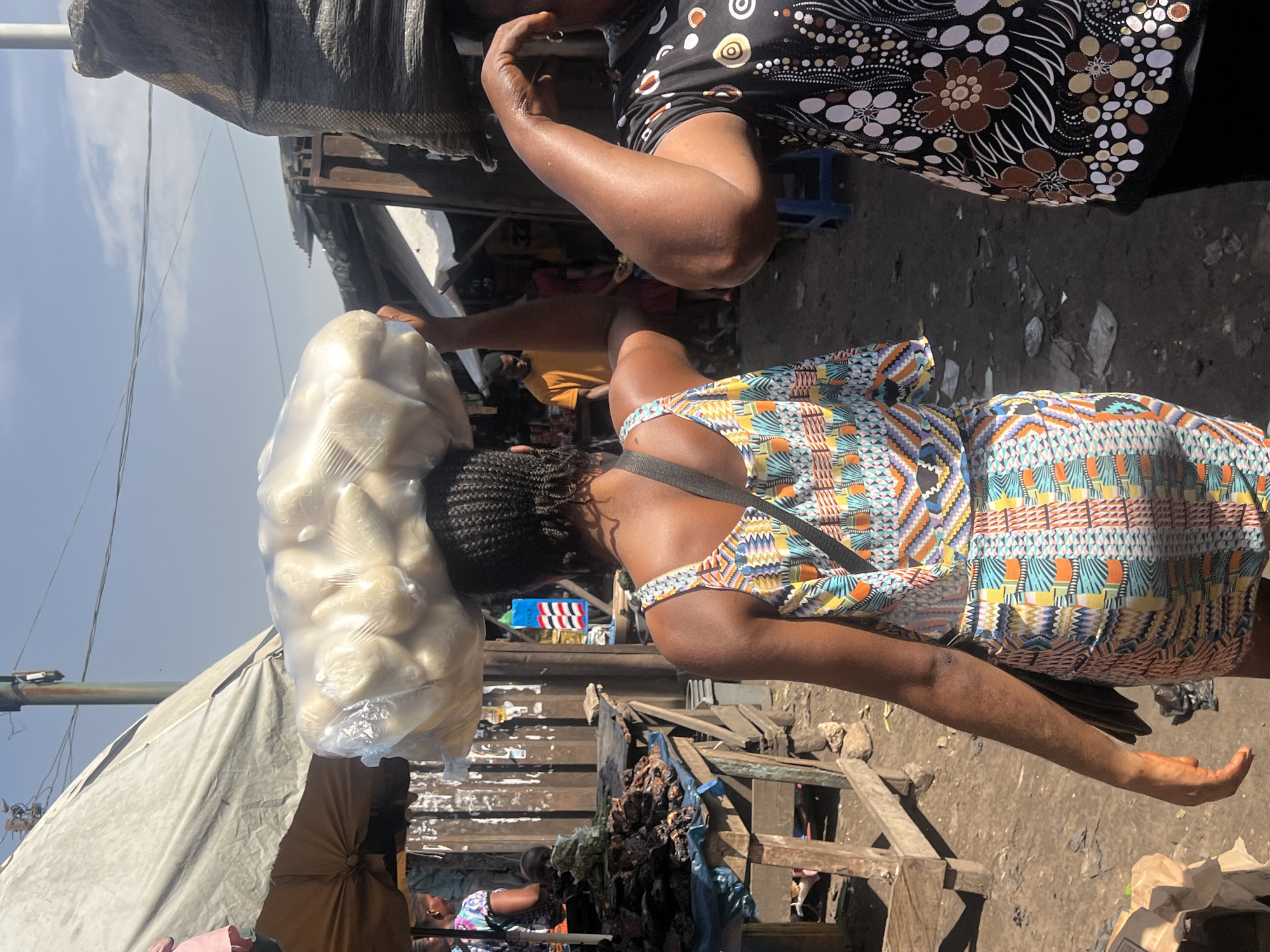 In Nkpor, Nigeria, a woman in a colorful patterned dress carries a large plastic bag of what appears to be food items on her head while walking on a dirt street in what appears to be a market. Another woman in a floral patterned shirt is standing nearby. Wooden structures and tables are present on either side of the street. There is a variety of other people and goods visible in the background.