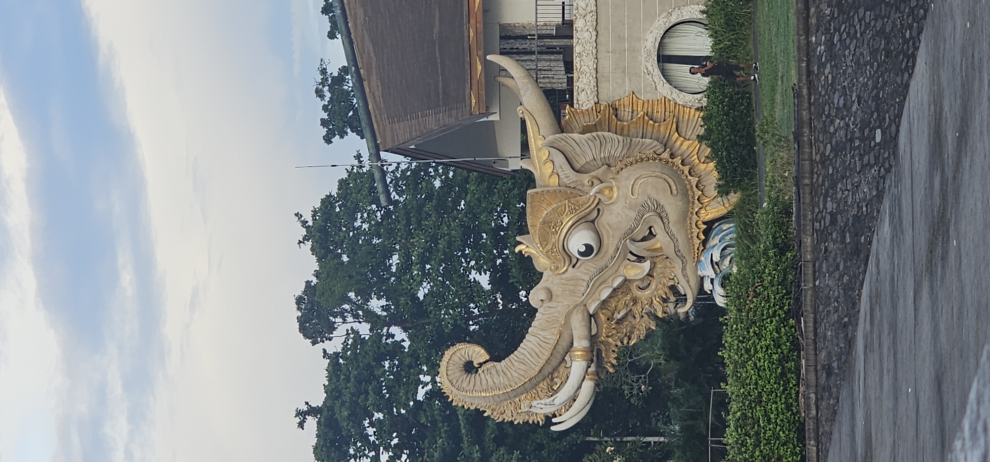 When correctly oriented, the image presents a striking view of a large, ornate statue resembling a mythical elephant-like creature, dominating the central and left portions of the frame. The statue is primarily light beige or off-white, adorned with elaborate golden accents on its crown, mane, and decorative elements. It features a long, curled trunk, prominent white tusks, and large, intricately detailed ears, with a single, prominent eye looking forward. Blue, wave-like patterns are visible near its base, partially obscured by green hedges.
Behind the statue, lush green trees fill the mid-ground, suggesting a tropical environment. Above the trees, a light blue sky with scattered, elongated white clouds stretches across the top of the image. To the right of the statue, part of a building with a dark, possibly tiled, roof and light-colored walls is visible. A distinctive circular window with white blinds is set into the building's facade. A man, wearing a dark shirt, dark shorts, and white sneakers, stands near this window, looking towards the left.
In the foreground, a neatly trimmed green hedge runs along the base of the statue and the building. To the right of the hedge, a textured stone wall or paving abuts a darker, smoother paved surface, possibly a road or pathway, which appears slightly wet.
The scene is captured during the daytime, under a clear sky with some cloud cover. The intricate artistry of the statue strongly indicates a cultural context typical of Southeast Asia, consistent with the specified location of Denpasar, Indonesia. There is no specific activity taking place beyond the man standing, and no visible text in the image.