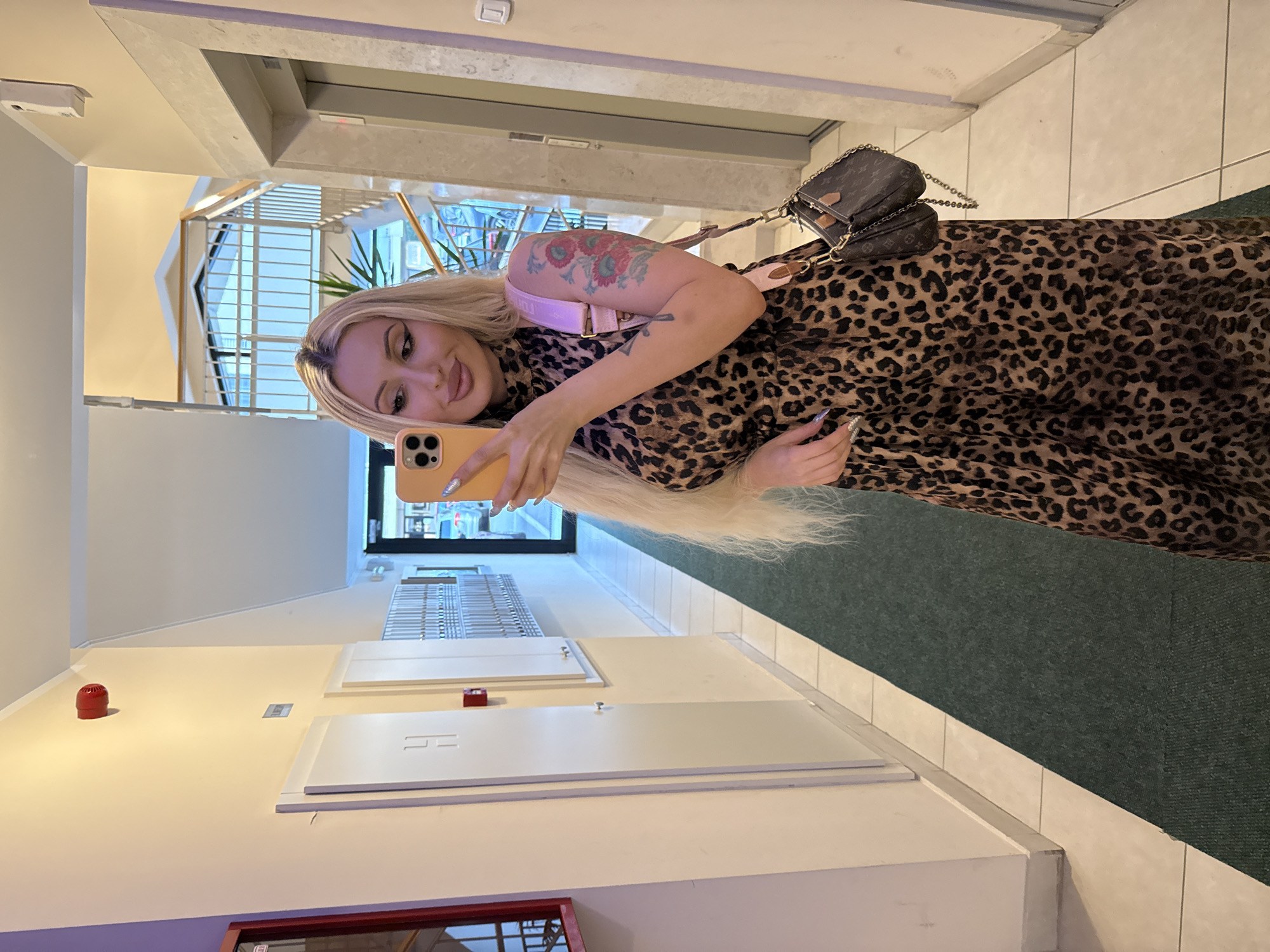 A blonde woman is taking a mirror selfie in a well-lit indoor hallway. She is wearing a long, leopard-print dress and a small designer handbag. Her left arm, which is visible, has a tattoo of red and pink flowers. She is holding an orange smartphone in her right hand, angled to capture her reflection. The hallway features a green carpet runner, cream-colored walls, and several doors on the left side, one of which has a red fire alarm mounted on it. To the right, there is an elevator and a staircase with a modern railing visible in the background. The overall environment suggests an apartment building or a similar residential complex.