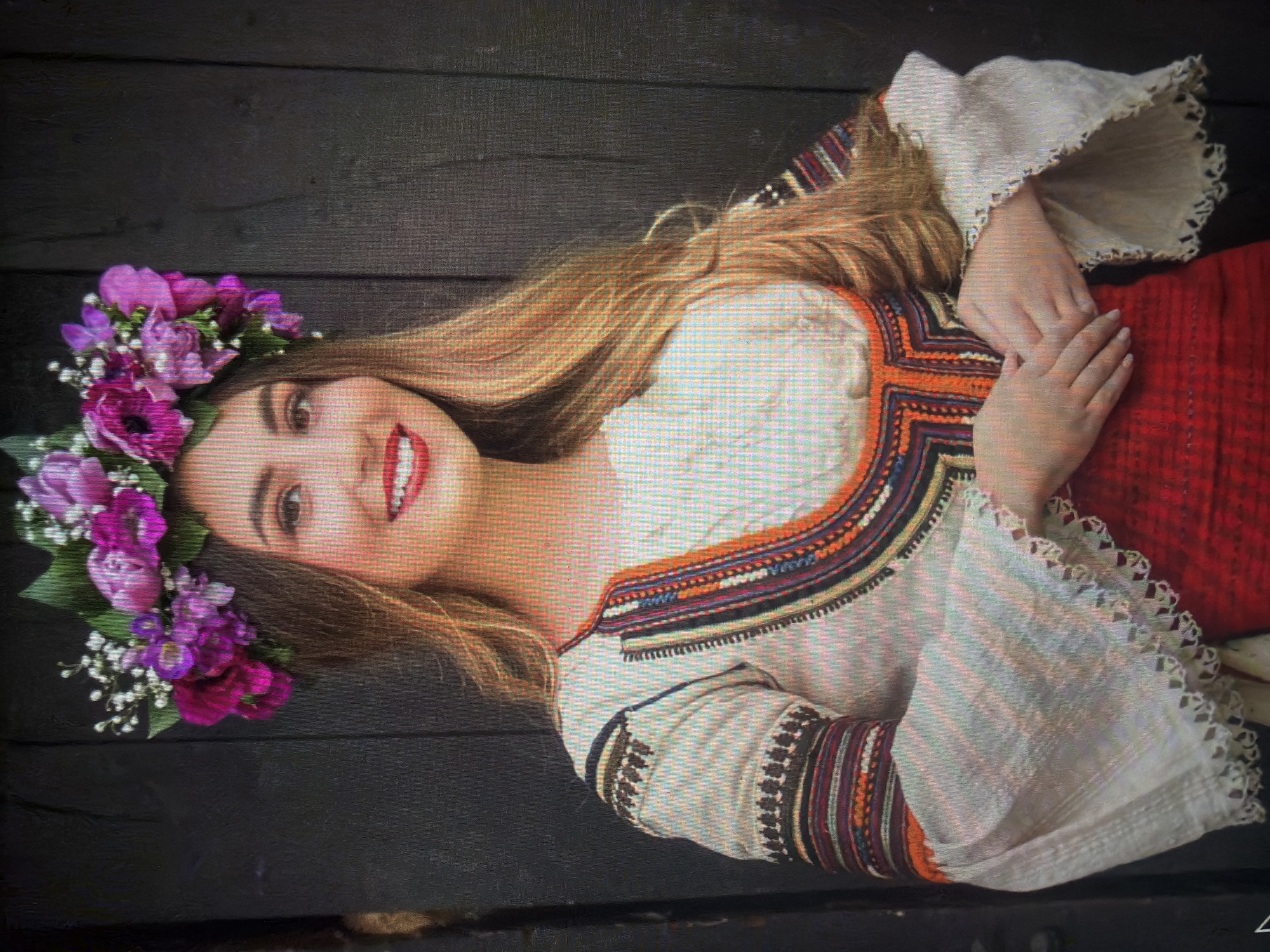 A young woman, appearing to be in her late teens or early twenties, is depicted in a mid-shot portrait. She has long, light brown hair styled with a slight wave, extending over her shoulders. Her complexion is fair, and she is smiling, revealing her teeth, with red lipstick applied to her lips. She wears a floral crown adorned with purple, magenta, and white flowers, interspersed with green leaves. Her attire consists of a white, traditional folk blouse with intricate multi-colored embroidery in shades of orange, black, blue, white, red, and yellow around the neckline and on the sleeves. The sleeves feature wide, bell-shaped cuffs with a distinctive scalloped, embroidered edge. A portion of a red skirt or lower garment is visible from her waist down. Her hands are clasped together in front of her lower torso. The background consists of a dark, vertically planked wooden surface, indicating a wall or door. The scene is identified as being in the city of Shumen, Bulgaria.