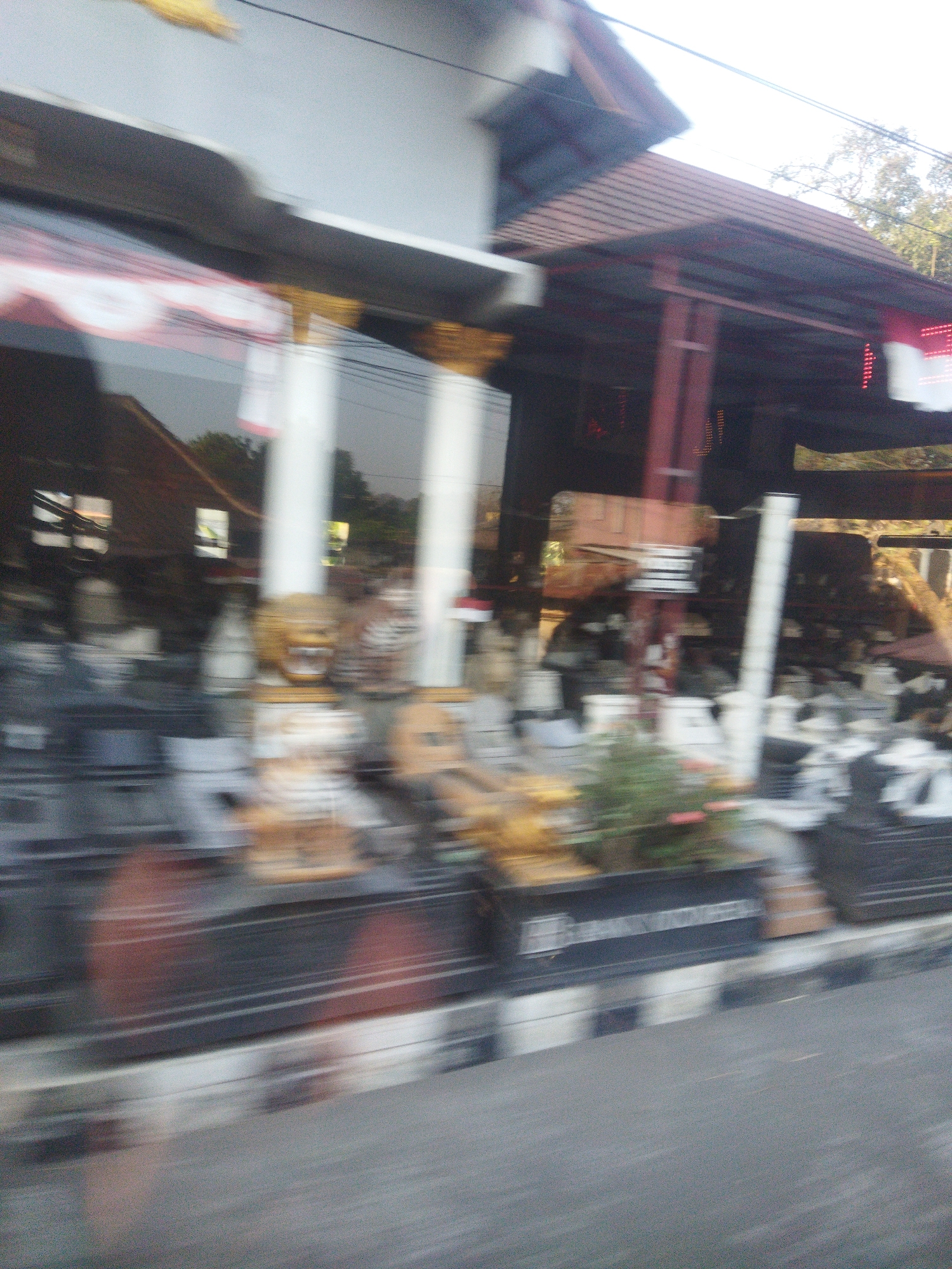 The image captures a blurred street view of a building with a dark roof and maroon pillars, possibly in Singodutan, Indonesia. The building appears to house a shop or establishment, with its front displaying various items, including statues or decorative objects. A large window reflects the surroundings, showing trees and parts of other buildings. A striped curb is visible along the street, indicating a pedestrian walkway or roadside. The overall impression is of a busy urban environment, with the motion blur suggesting the photograph was taken from a moving vehicle.