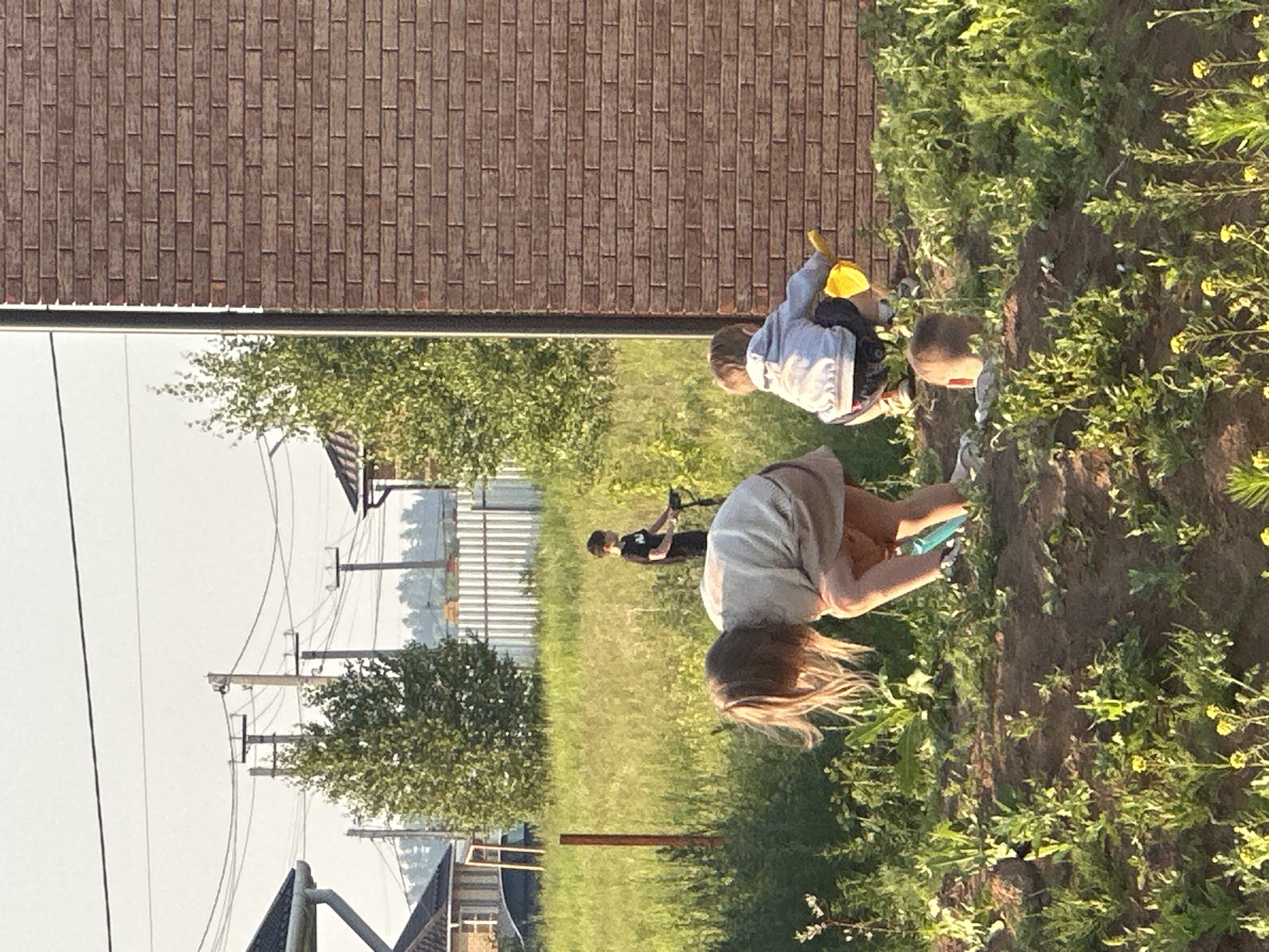 The image shows a backyard scene in Estachi, Russia, with four children engaged in outdoor activities. In the foreground, a young girl with long blonde hair is bent over, appearing to dig in the dirt with a blue tool. To her right, another child, wearing a grey hoodie and dark shorts, is crouched and holding a yellow toy shovel. A third child, a young boy wearing a black t-shirt, is visible further in the background, standing on a scooter. A fourth child's head is visible near the bottom of the frame, appearing to be lying down or crawling in the dirt. The environment consists of a garden area with green plants and yellow wildflowers, a dirt path, and a brick wall on the right side of the frame. In the background, there are trees, a white fence, power lines, and houses, suggesting a residential area. The lighting indicates it is daytime, likely late afternoon or early evening due to the warm light.
