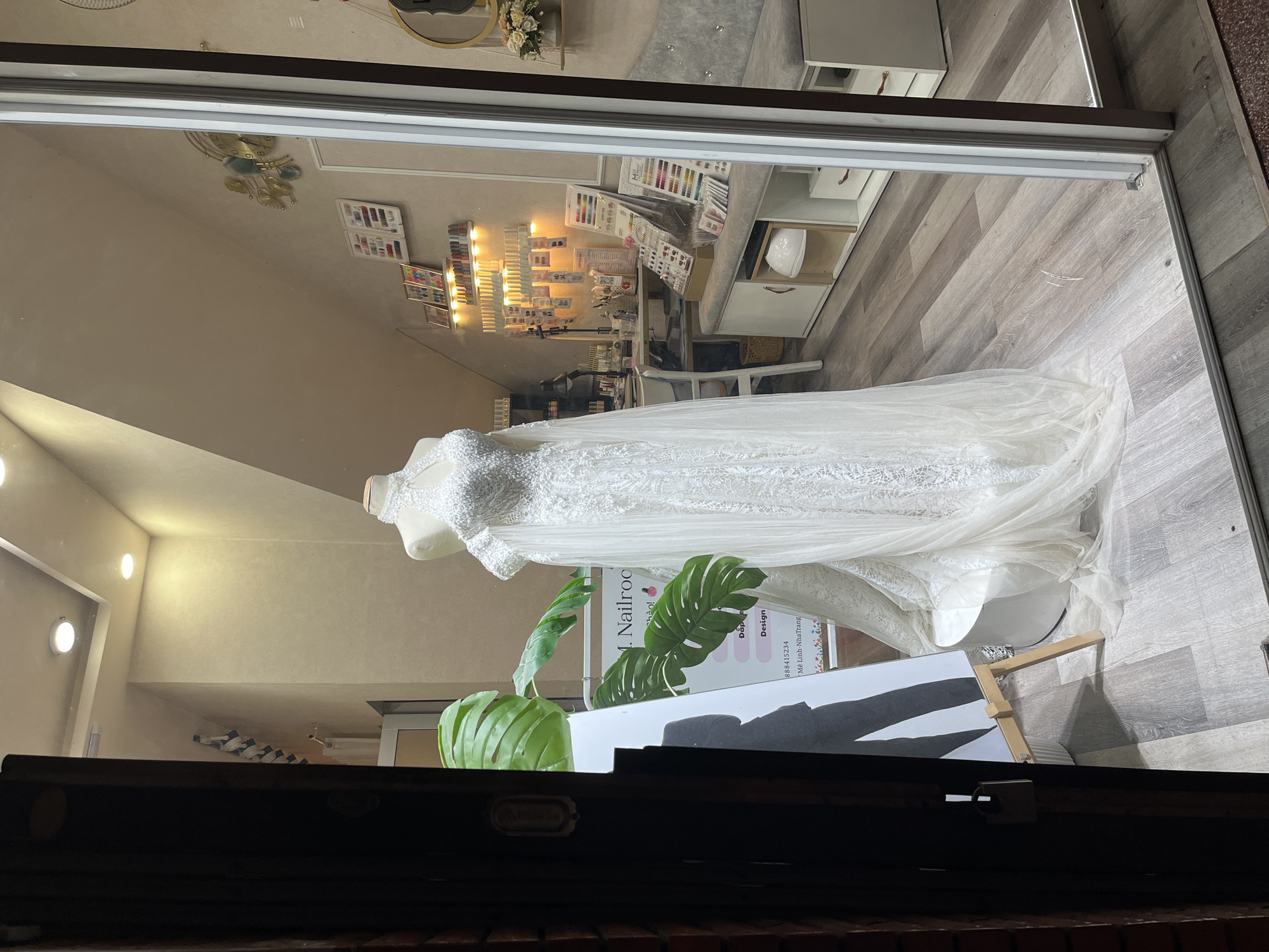 The image depicts a wedding dress boutique or salon window display in Nha Trang, Vietnam. A white wedding dress is prominently displayed on a mannequin. Behind the dress, the interior of the shop is visible, featuring shelves stocked with nail polish bottles, nail art supplies, and a work desk with a lamp and various tools, suggesting the business also offers nail services. A large green monstera plant is placed to the left of the wedding dress. A sign with Vietnamese text, partially obscured by the plant, includes the words "M. Nailroo" and "Design". Further in the background, a light-colored bench with tufted upholstery and a round mirror with decorative accents are visible. The floor is made of light-colored wood-look laminate. The overall scene is brightly lit and orderly, showcasing the merchandise and services offered by the establishment.