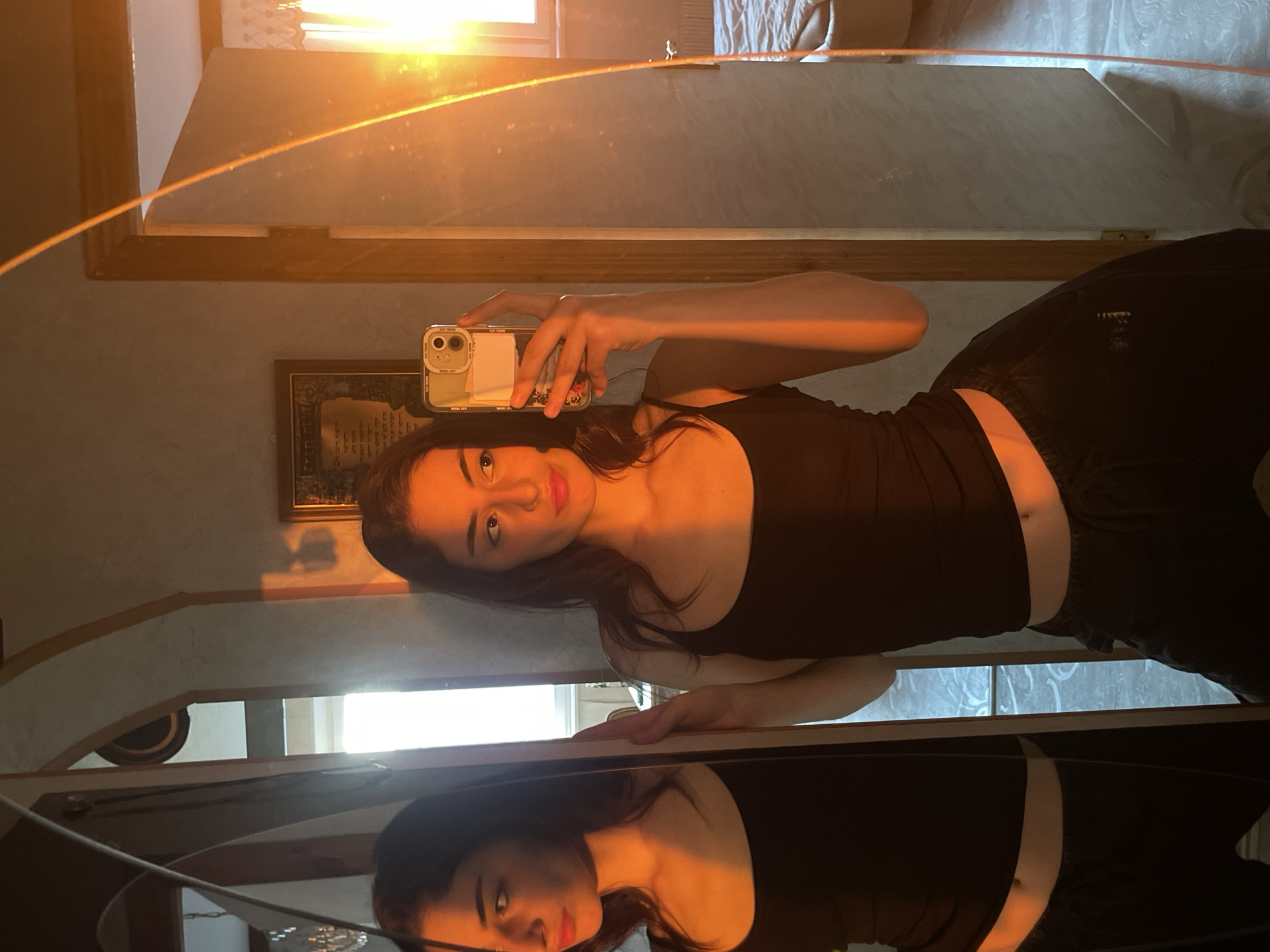 A woman is taking a selfie in a mirror. She is wearing a black tank top and black pants. The background shows parts of a room with a bed and other furniture. A framed picture hangs on the wall. There is no verifiable indication of the location being Moscow, Russia.