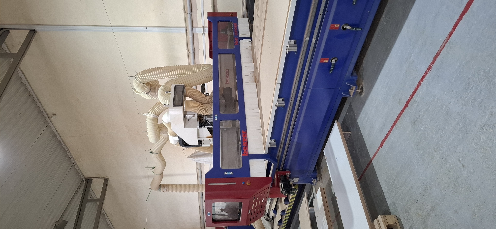 The image depicts an industrial setting, likely a workshop or factory, in Aktau, Kazakhstan. A large, blue and red CNC (Computer Numerical Control) cutting machine, branded "Beaver," is the central focus. The machine features a long bed for material placement and multiple flexible, cream-colored hoses extending from an overhead system, presumably for dust collection. The surrounding environment consists of a concrete floor marked with a red line, and stacks of flat, white materials, likely wood or composite boards, are visible in the foreground. The walls are a pale, textured material, and the ceiling appears to be corrugated metal with exposed beams. A person can be seen reflected in a window on the left side of the machine. The overall impression is one of a functional, active production facility.
