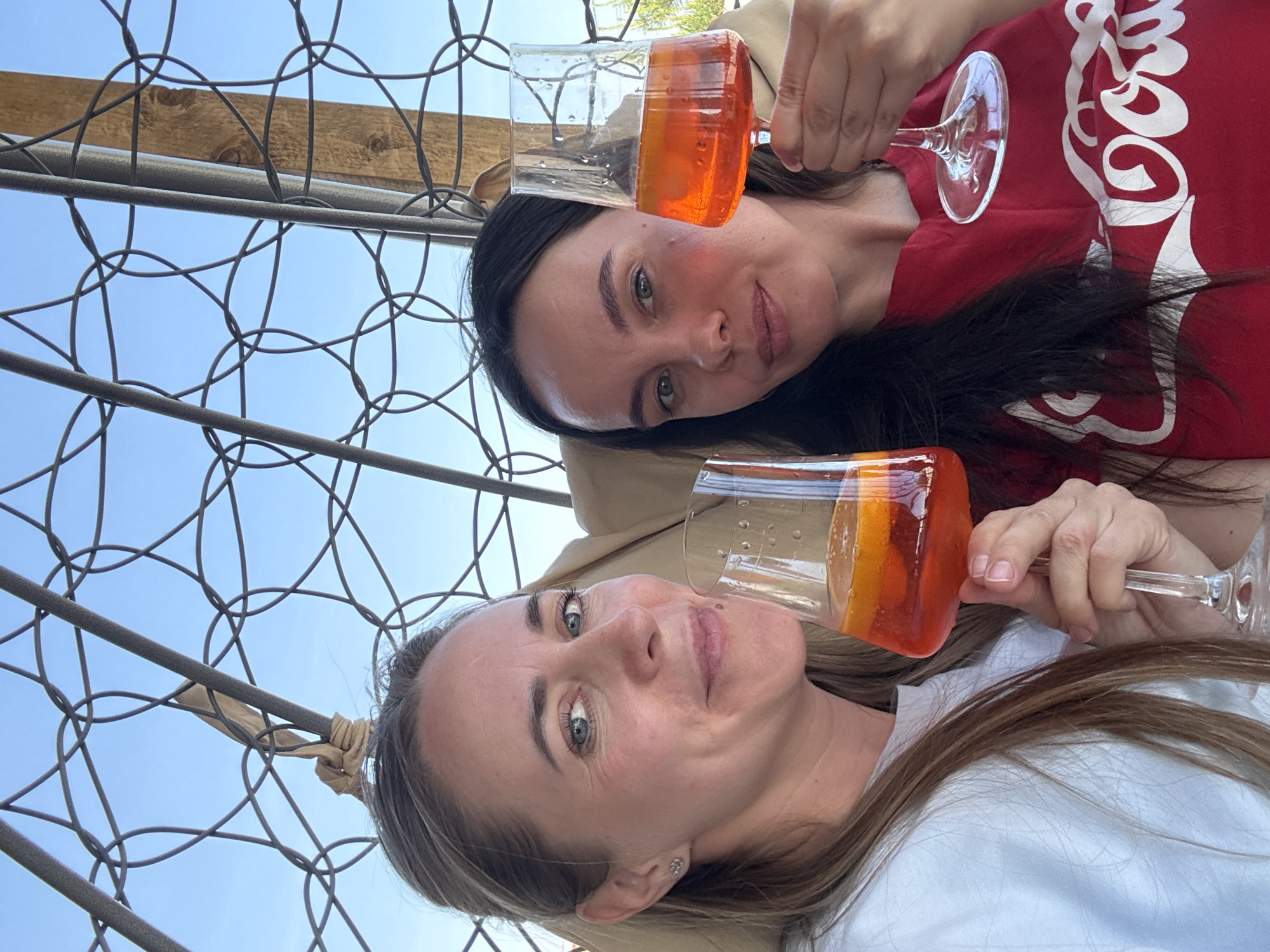 Two women pose in a close-up shot, each holding a glass of an orange-colored beverage. The woman at the bottom of the frame has blonde hair and wears a white shirt, while the woman at the top has dark hair and wears a red shirt with white lettering. The photo appears to be taken from a low angle, looking up through a lattice-like structure of gray metal. Blue sky is visible through the metal framework. The image was taken in the city of Chaikovsky, Russia.