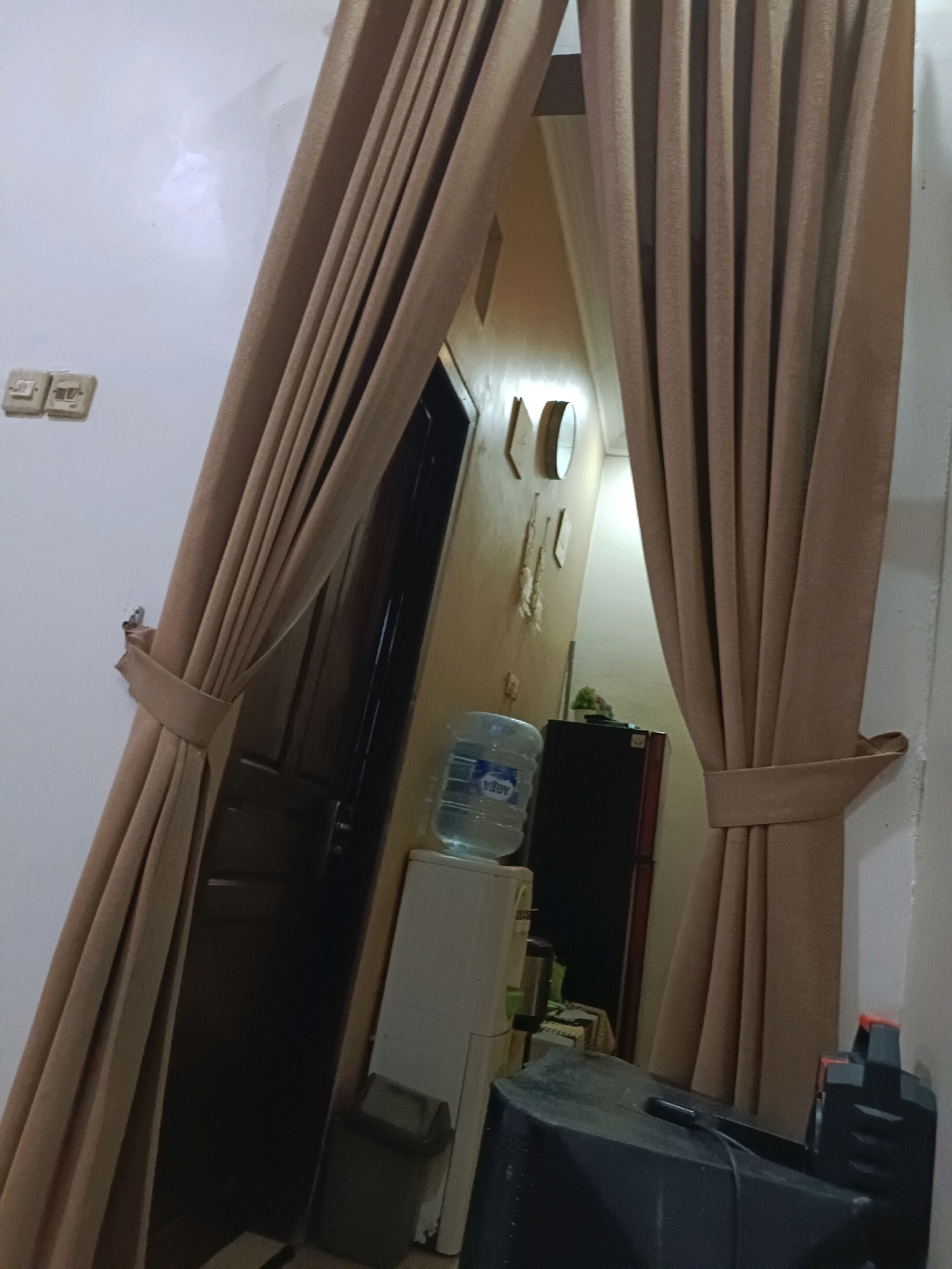 The image depicts an indoor scene, likely a residential area in Rancang Kencono, Indonesia. The foreground is dominated by large, beige curtains that hang from what appears to be a curtain rod. These curtains are partially drawn, revealing a glimpse of a doorway and a narrow hallway beyond.
To the left of the curtains, a white wall is visible with two electrical outlets mounted on it. Further into the hallway, a dark-colored door is partially open. Adjacent to the door, a white water dispenser with a large plastic bottle of water on top is present. Below the water dispenser, a dark-colored trash bin can be seen.
The hallway itself is illuminated, with a warm yellow hue emanating from the wall on the right side. On this wall, there are several decorative items, including what appear to be small framed pictures or plaques, and a circular mirror. Further down the hallway, a dark refrigerator is visible, and to the right of that, a portion of a beige curtain is seen again, suggesting the hallway leads to another room.
In the lower right corner of the image, a large black object, possibly a speaker or a piece of electronic equipment, is visible. Its surface is matte black and appears to be slightly dusty. The lighting in the scene is dim, with the primary light source likely coming from the hallway. The overall impression is one of a lived-in, domestic environment. There are no people visible in the image.
