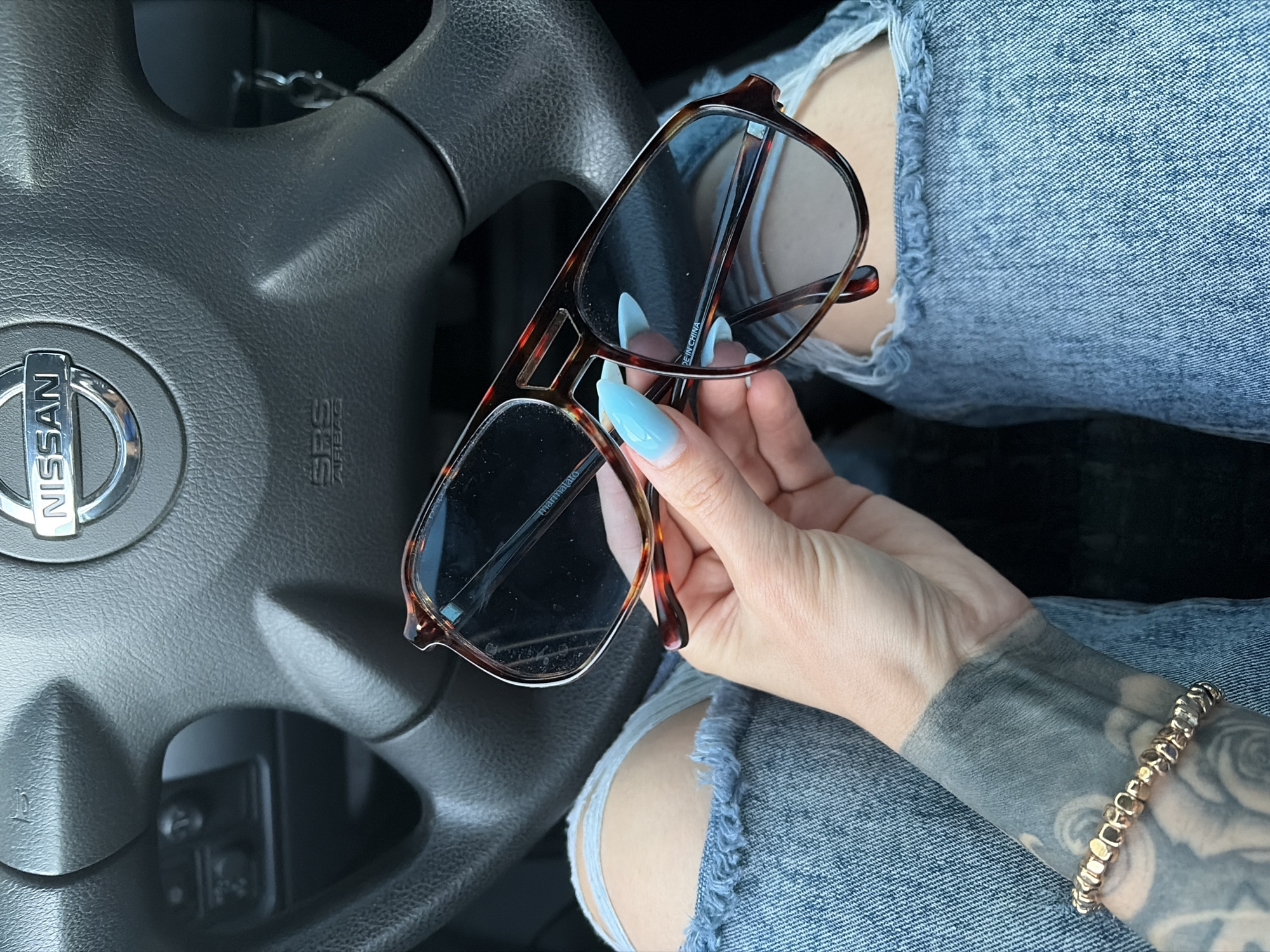 A hand with light blue fingernails holds a pair of tortoiseshell glasses over the driver's side of a car. The steering wheel with a Nissan logo is visible to the left. The person is wearing ripped denim jeans. The arm of the person, visible on the right, has a tattoo of roses and a beaded bracelet. The lighting suggests it is daytime, and no weather conditions are apparent. The scene appears to be inside a car, likely parked. The visible text on the glasses is "marmalato" and "MADE IN CHINA."