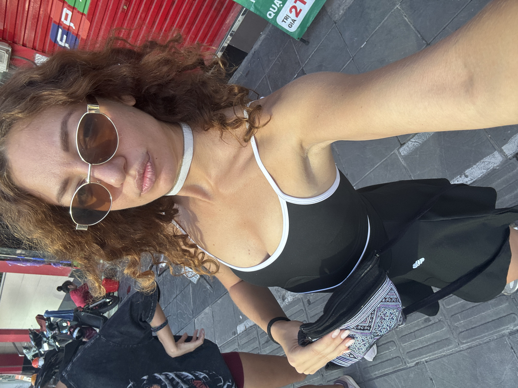 In a city street scene in Hanoi, Vietnam, a woman with curly brown hair and sunglasses takes a selfie. She is wearing a black and white-trimmed tank top, a white choker necklace, and a black skirt. She is holding a black purse with a patterned front. In the background, motorcycles are parked, and a person in a red shirt is washing a bucket. A red shuttered building with signage is visible to the right. The ground is paved with grey tiles.