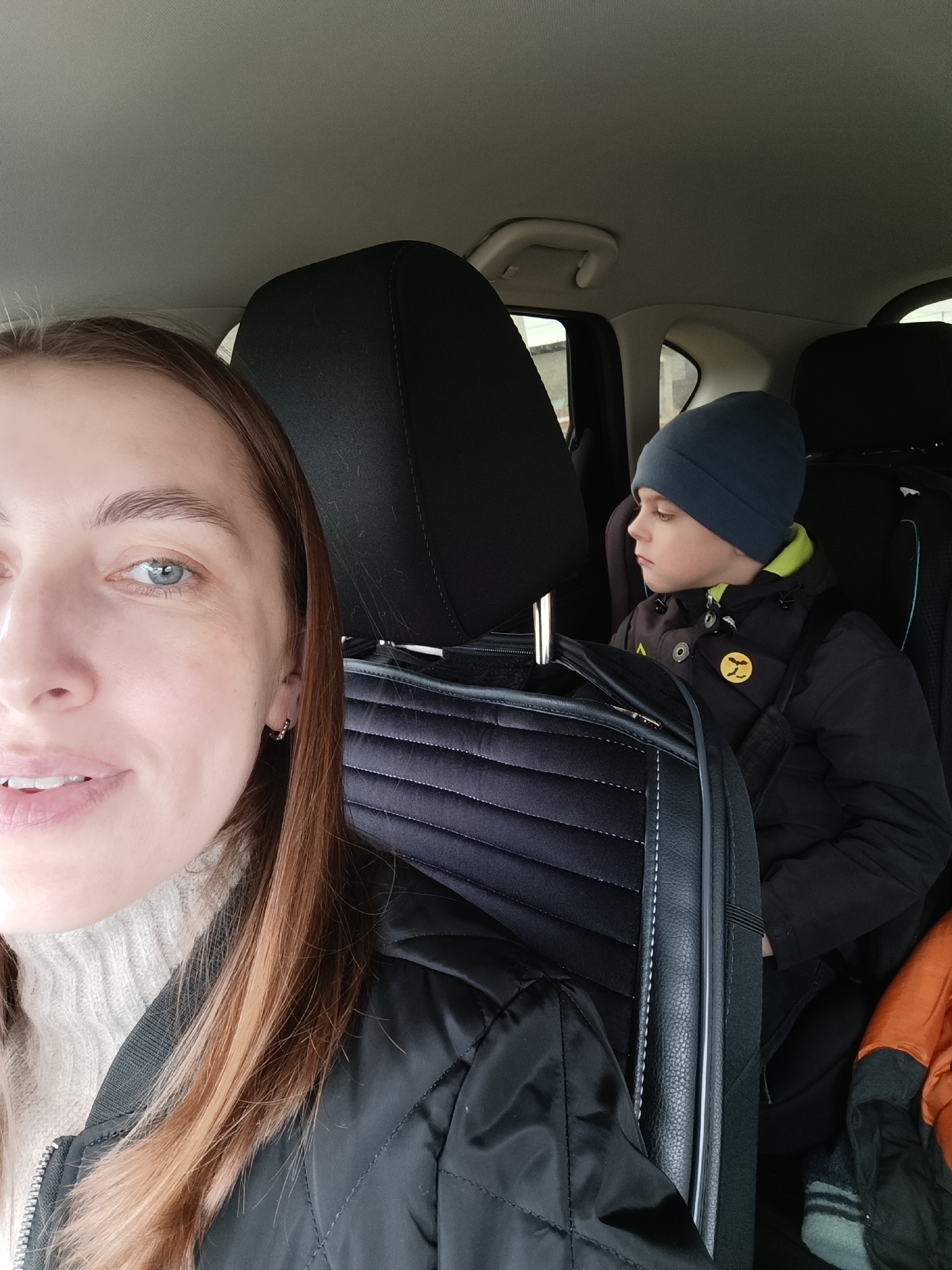 A woman and a child are in a car. The woman is in the front seat, and the child is in the back. The woman is wearing a light-colored sweater and a black jacket. The child is wearing a dark beanie, a dark jacket with lime green accents, and has a circular yellow patch on the jacket. A black quilted bag is between the woman and the child. The background shows car interiors. Based on the visible environment and objects in the image, determining the specific location within the city of Chaikovsky, Russia, is impossible.