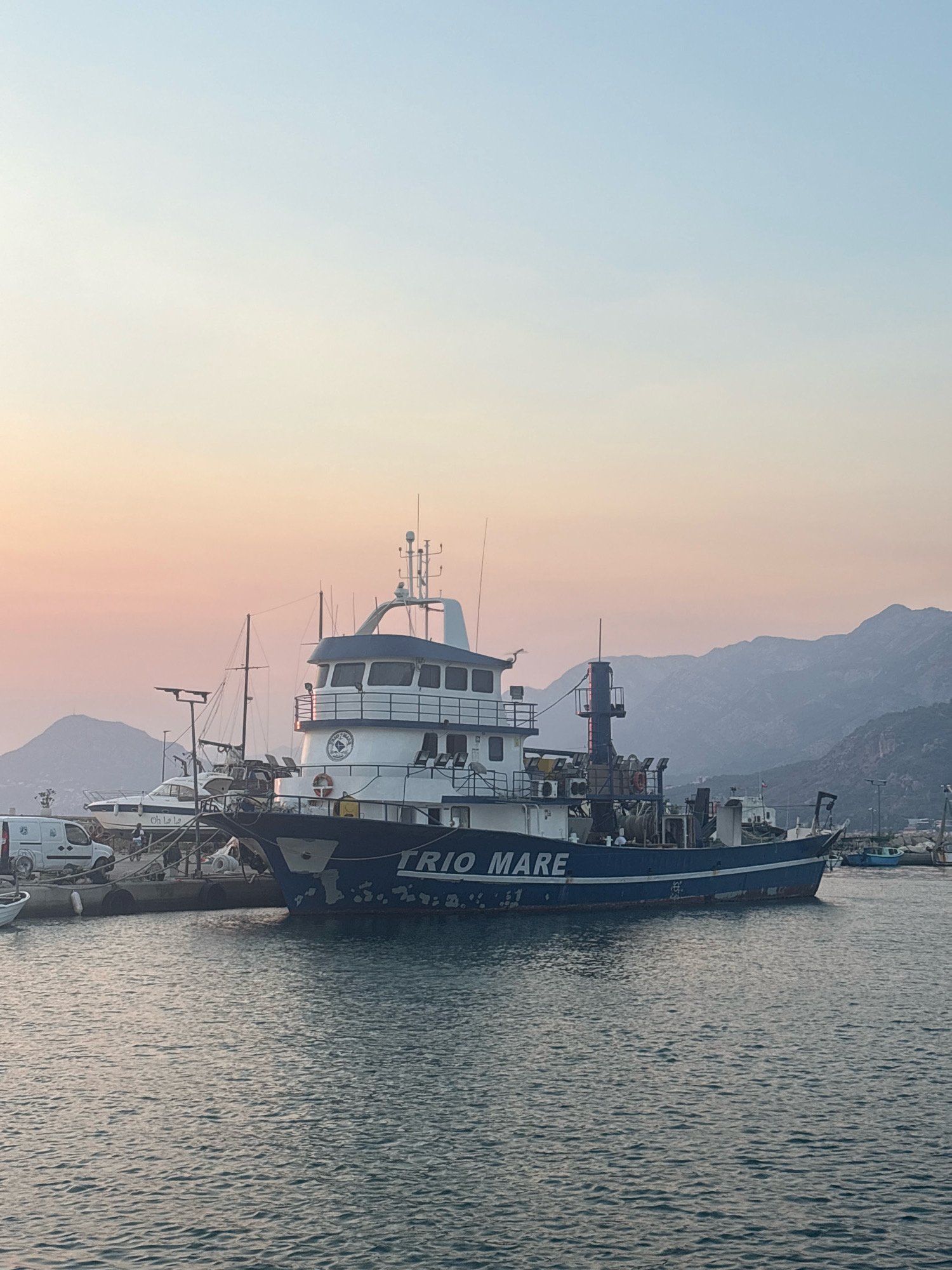 This image shows a fishing vessel named "TRIO MARE" docked in the port of Bar, Montenegro. The vessel is blue and white with "TRIO MARE" written in white lettering along its side. It is moored alongside other boats, including a white yacht with the name "Oh La La" visible. In the background, there are mountains and a hazy sky with soft pink and blue hues, suggesting either sunrise or sunset. A white van is parked on the dock near the boats. The water in the foreground is calm with slight ripples.