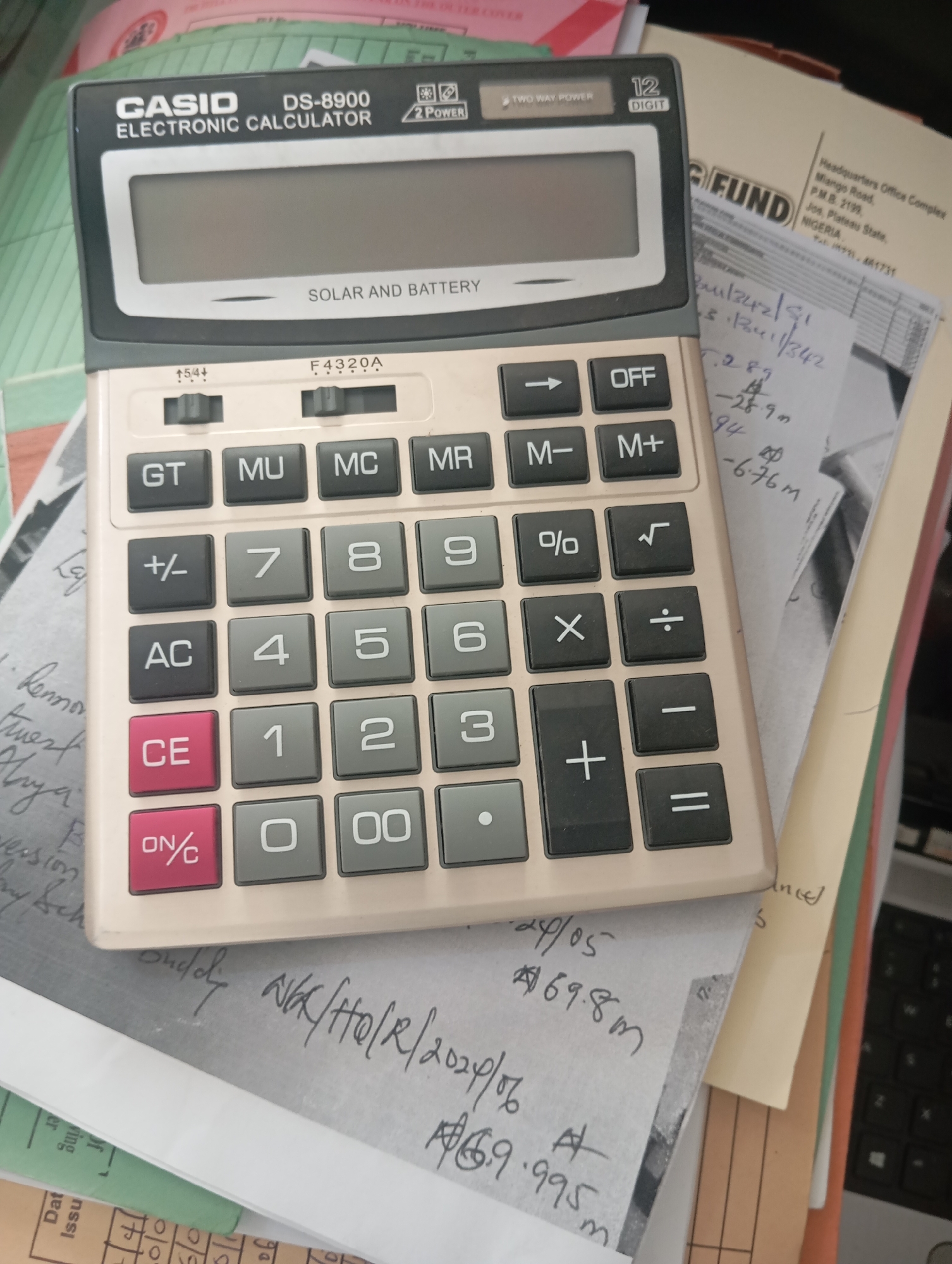 The image shows a CASIO DS-8900 Electronic Calculator placed on top of a pile of documents. The calculator is beige and has a solar panel at the top. It displays "SOLAR AND BATTERY" below the screen. The keyboard has numerical buttons from 0-9, function buttons like GT, MU, MC, MR, +/-, AC, CE, ON/C, and operational buttons like +, -, x, ÷, =, %, √, OFF, and an arrow button.
Beneath the calculator, several handwritten notes and official documents are visible. One document has "Headquarters Office Complex, P.M.B. 2199, Jos, Plateau State, NIGERIA" printed on it, indicating the location as Jos, Nigeria. Another handwritten note contains a series of numbers and symbols, some appearing to be calculations. There are also some green and yellow folders or papers. The overall environment appears to be an indoor setting, possibly an office, with papers and a calculator scattered on a surface. There are no people visible in the image.