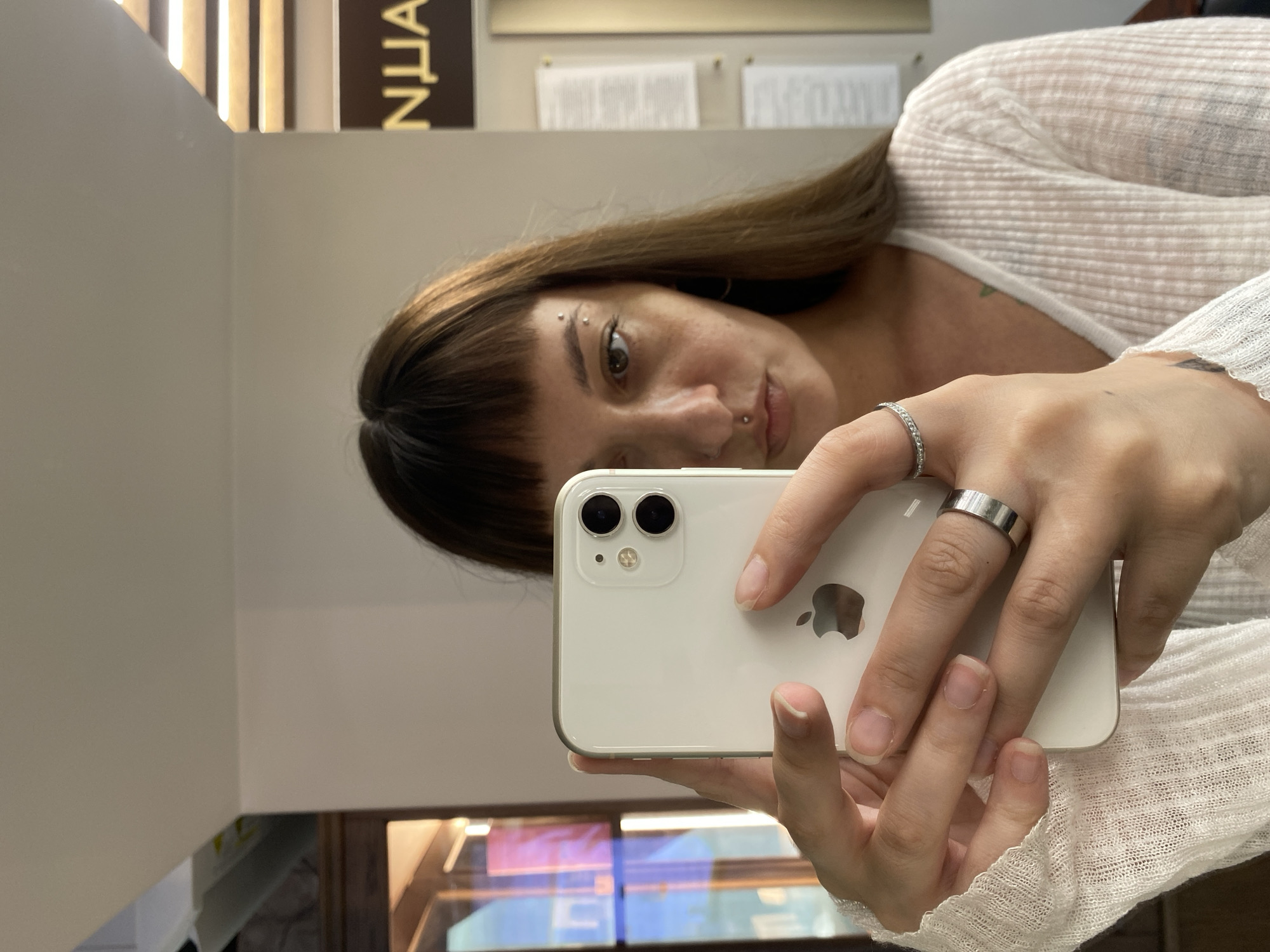 In a well-lit interior, likely a private residence or small office in Votkinsk, Russia, a young woman is captured taking a selfie with her iPhone 11. The phone is held horizontally, with the rear camera facing the subject. The woman has long, dark brown hair with bangs and several piercings on her face, including one above her left eyebrow and another on her lower lip. She is wearing a thin, white ribbed long-sleeved shirt. Her hands, adorned with two rings, are visible holding the phone. The background shows a white wall, part of a wooden cabinet on the left, and a sign with the Cyrillic letters "NUA" on the right. The overall impression is of a casual, personal moment documented in an everyday setting.