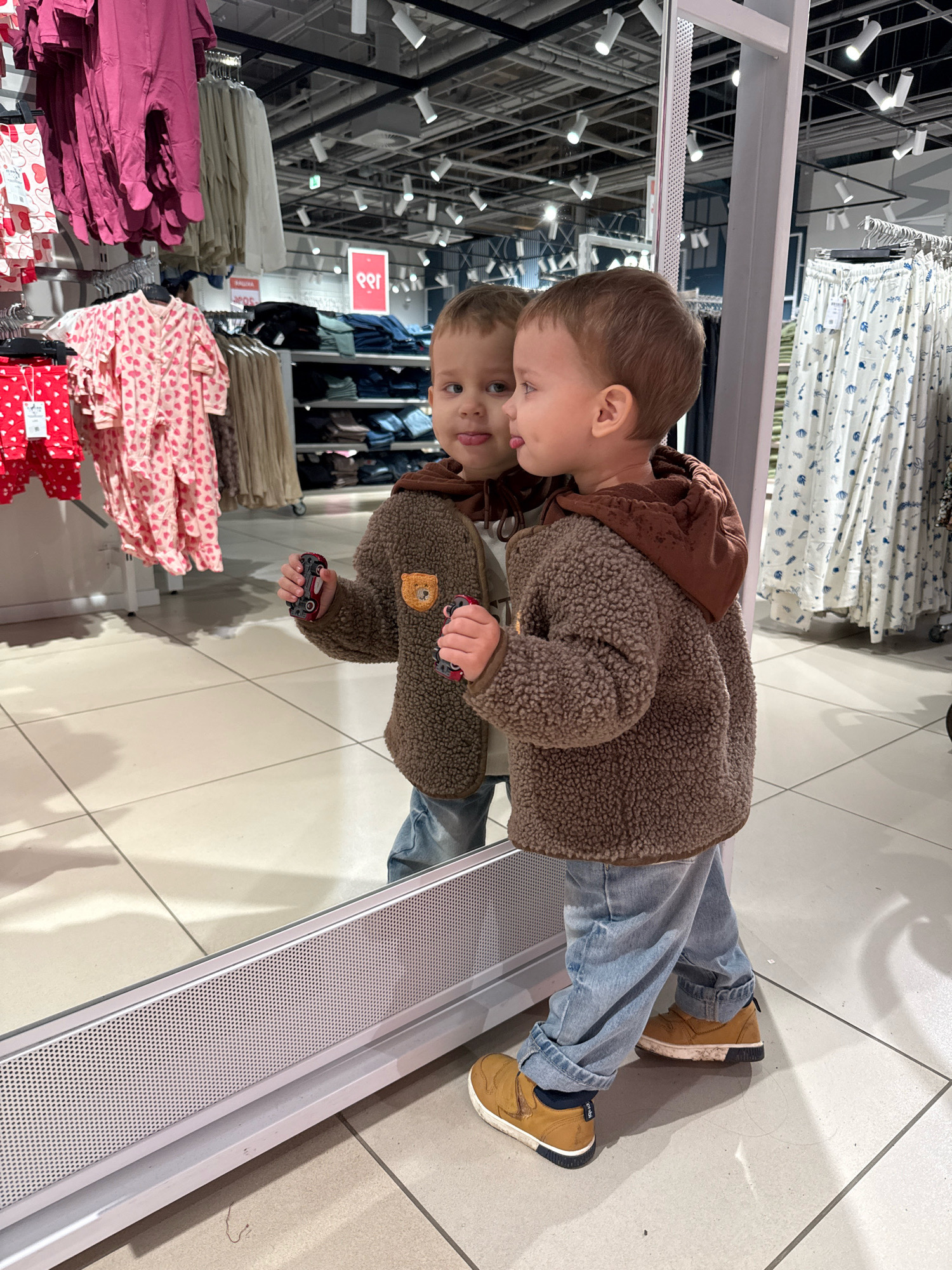 In the city of Zhukovo, Russia, a young boy stands in front of a large mirror in a retail store, likely a clothing store. He is wearing a brown fluffy jacket with a hood, light blue jeans, and brown slip-on shoes. The boy is holding two small toy cars, one in each hand. His reflection in the mirror shows him sticking his tongue out. The background of the image reveals racks of clothing, including pink onesies with hearts and white patterned pants. The lighting appears to be from overhead fluorescent lights, typical of a commercial setting. The floor is tiled with light-colored squares.