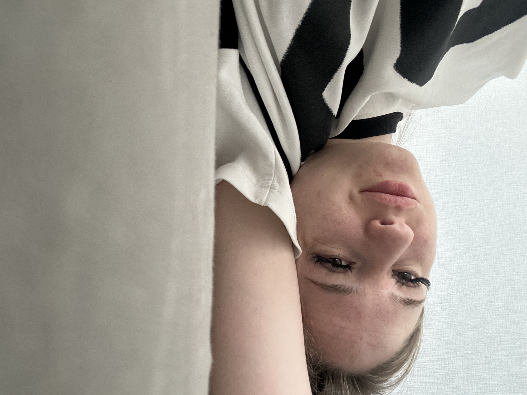The image displays a close-up, inverted view of a young adult female, suggesting she is lying down and looking up towards the camera. Her face, featuring light skin and light hair framing her temples, dominates the right side of the frame. She has distinct eyelashes, and her lips are slightly parted, conveying a calm or relaxed expression.
She is wearing a black and white top with bold, abstract black patterns on a white fabric. Her right arm, bent at the elbow, is visible alongside her head and shoulders. The background on the left consists of a solid, light grey surface, possibly a wall or fabric. On the right, behind her head, a lighter, subtly textured surface, likely a ceiling or another wall, completes the indoor setting. The lighting is soft and ambient, offering no specific cues for the time of day. There is no visible text.