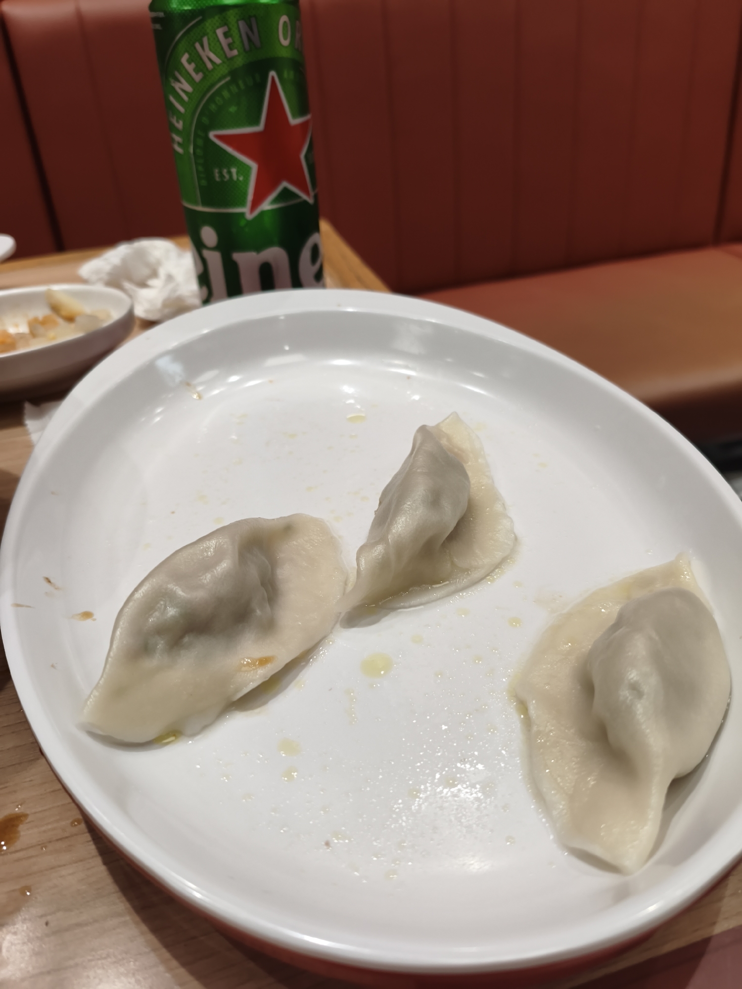 A white plate with three dumplings sits on a wooden table. A can of Heineken beer is to the left of the plate. In the background, a red padded seat is visible. The setting is likely a restaurant in Mudanjiang, China.