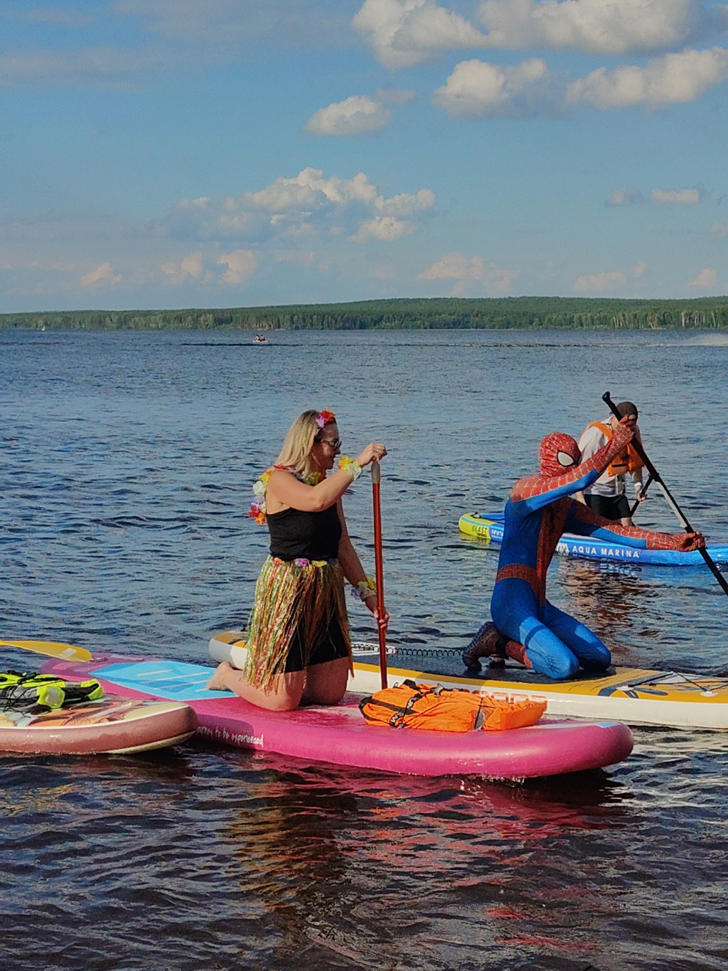 Three people are paddleboarding on a body of water under a partly cloudy sky. In the foreground, a woman wearing a black tank top, a hula skirt, and a flower lei kneels on a pink paddleboard. She holds a paddle in her right hand. To her right, a person dressed in a Spider-Man costume kneels on a yellow and blue paddleboard, holding a paddle. Behind the Spider-Man figure, another person wearing an orange life vest and a hat paddles a blue and yellow paddleboard. The background shows a treeline and a body of water with a few small boats in the distance.