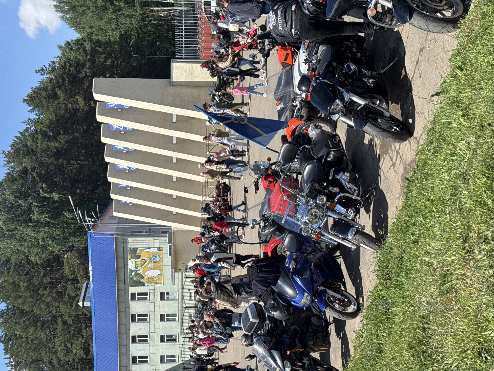 A wide-angle, eye-level photograph depicts a gathering of people and motorcycles in Chaikovsky, Russia, beside a multi-story building with a blue roof and a large mural.
The scene is arranged so that the building is situated on the left-hand side, partially obscured by dense green trees. The mural is visible and is a colorful representation of hockey players. In front of the building, a crowd of people are gathered, some standing and some seated.
To the right of the building, numerous motorcycles are parked in rows, with their front ends facing towards the viewer. The bikes are diverse in style and color, suggesting a motorcycle rally or gathering. A blue flag is visible amidst the crowd.
The environment includes a paved ground where the motorcycles and people are located, adjacent to a patch of grass on the right side of the frame. A large gray structure with a series of balconies is also visible and is associated with blue iconography. The sky is blue and clear. The photograph appears to be taken in daylight with natural lighting and shadows.