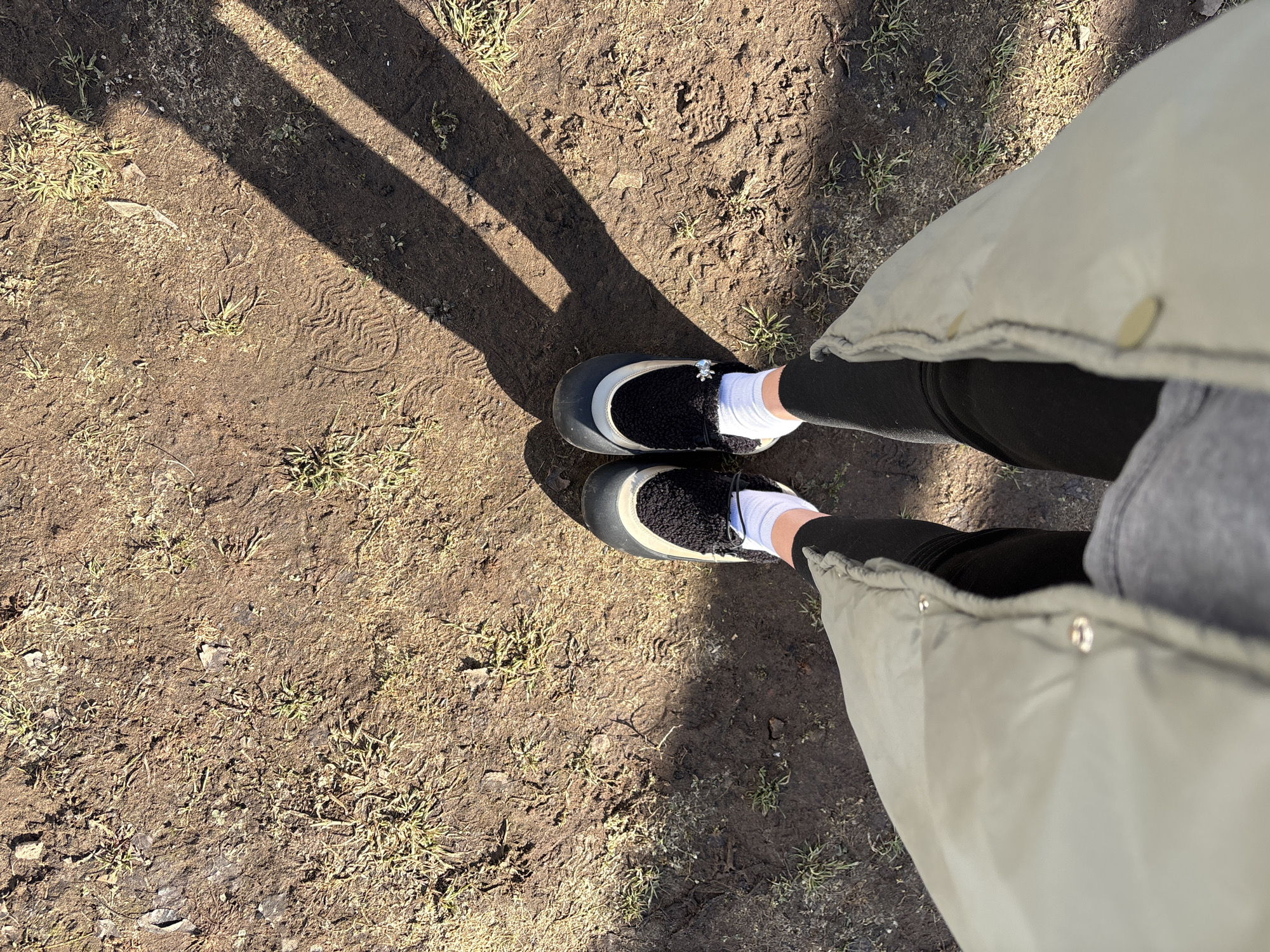 A top-down view depicts the lower body of an individual standing on a patch of uneven, disturbed brown soil mixed with sparse dry grass and small debris. The ground is illuminated by direct sunlight, casting several distinct, elongated parallel shadows from an unseen vertical structure onto the dirt surface, primarily on the right side of the frame. Faint shoe sole impressions are visible in the soil in multiple areas. The individual is wearing a light olive green or khaki quilted jacket with visible snap buttons, black leggings, and white ankle socks. Their feet are clad in black and off-white/grey slip-on shoes or boots, featuring a black faux fur or fleece lining. A small, metallic, spider-shaped charm is affixed to the upper right shoe. The scene is located in Votkinsk, Russia.