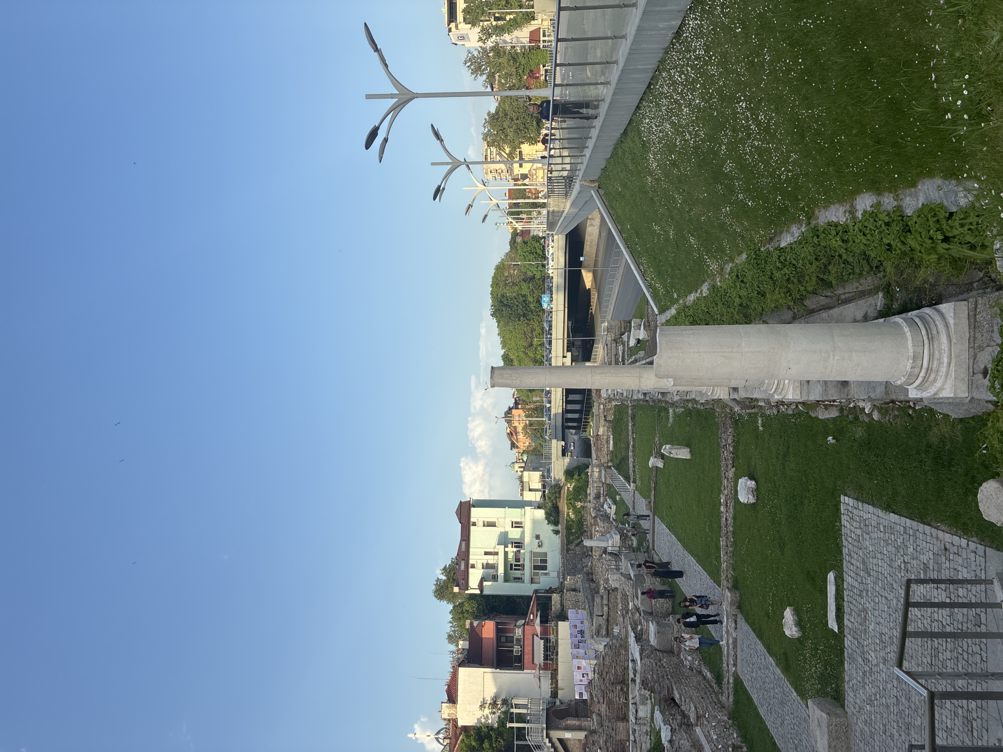 The image shows the Ancient Stadium of Philippopolis in Plovdiv, Bulgaria. The remains of the stadium are visible in the foreground. The remnants of the stadium walls and seating are made of stone and brick. Modern buildings and structures are visible in the background. Some people are walking and sitting on the steps and pathways around the site. There is a pedestrian overpass with flower-strewn grass visible overhead. The sky is blue.
