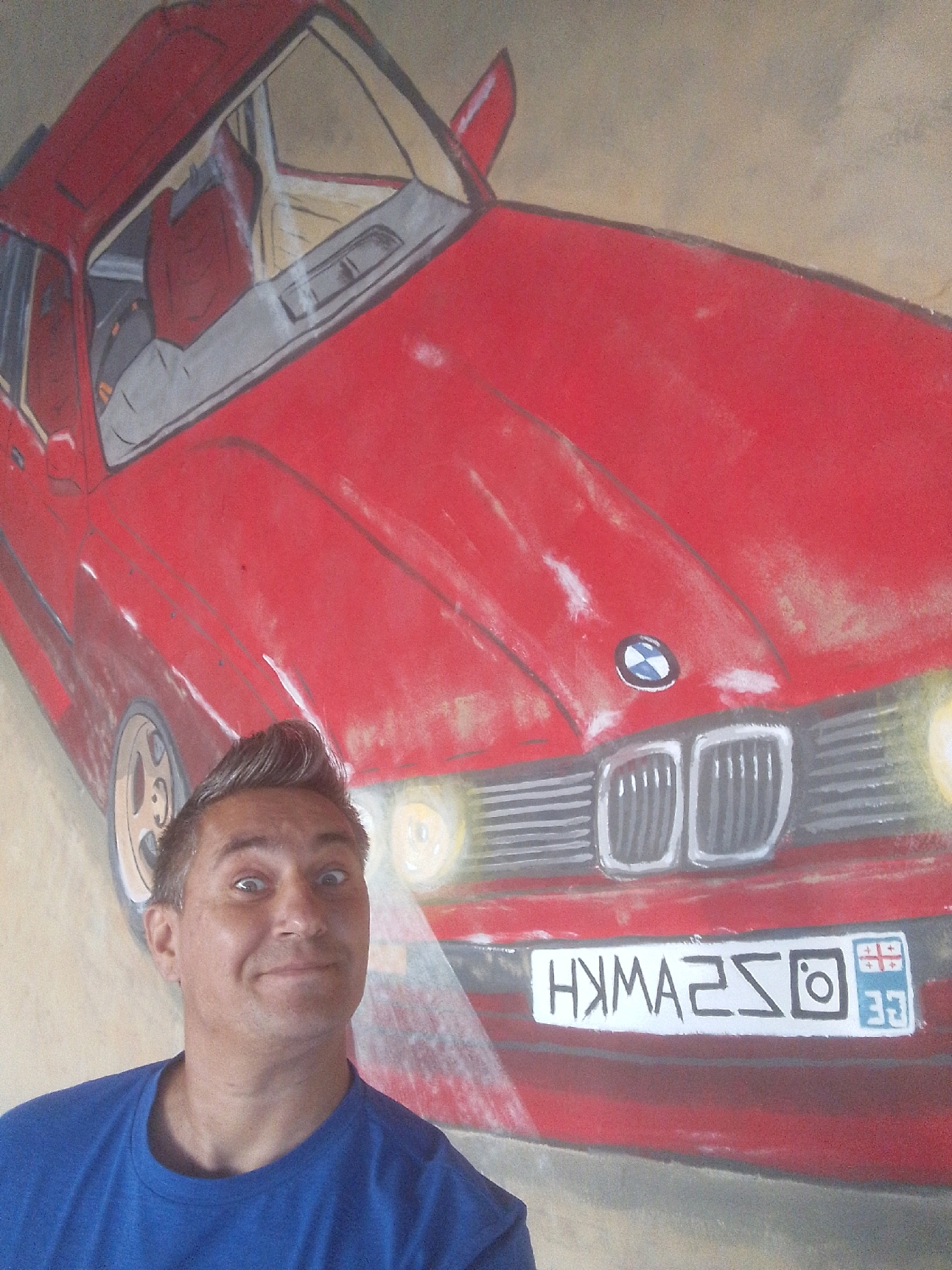 A man in a blue t-shirt is posing in front of a mural of a red car, possibly a BMW, painted on a wall. The mural is a close-up view of the car, showing its front grille, headlights, and a portion of the hood. The license plate on the mural features stylized characters and symbols, including an Instagram logo and what appears to be a flag with a cross design. The man's expression is one of surprise or jest, with wide eyes and a slight smile. The background appears to be a plain wall, suggesting this is an indoor setting, possibly in a studio or a building with murals. The overall scene captures a playful interaction between a person and street art.
