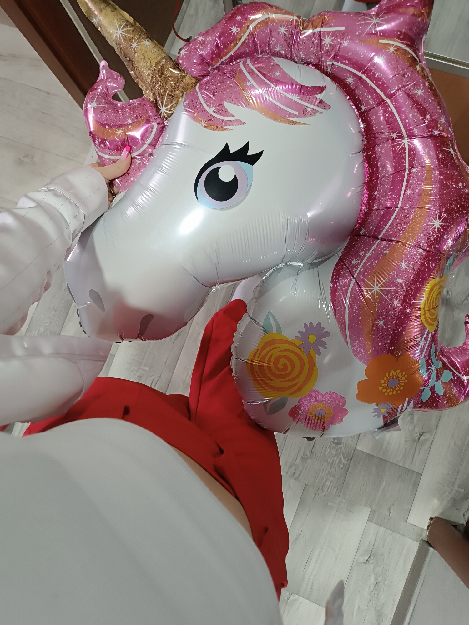 The image captures a large, inflated foil unicorn balloon held in an indoor environment. The balloon features a white head with a blue eye, black eyelashes, and a golden, glitter-accented horn. Its pink, glittery mane is adorned with white star patterns and orange stripes. The visible portion of the body is white with multicolored floral patterns in orange, yellow, and purple. A human hand, covered by a white long-sleeved garment and exhibiting pink-painted fingernails, grasps the unicorn's pink ear. A red garment is visible below the balloon, alongside a white garment or object. The flooring consists of light grey laminate wood planks. A partial view of a human foot is present on the right edge of the frame. Background elements include dark brown and light-colored surfaces. The location is an indoor space in Unknown, Unknown.