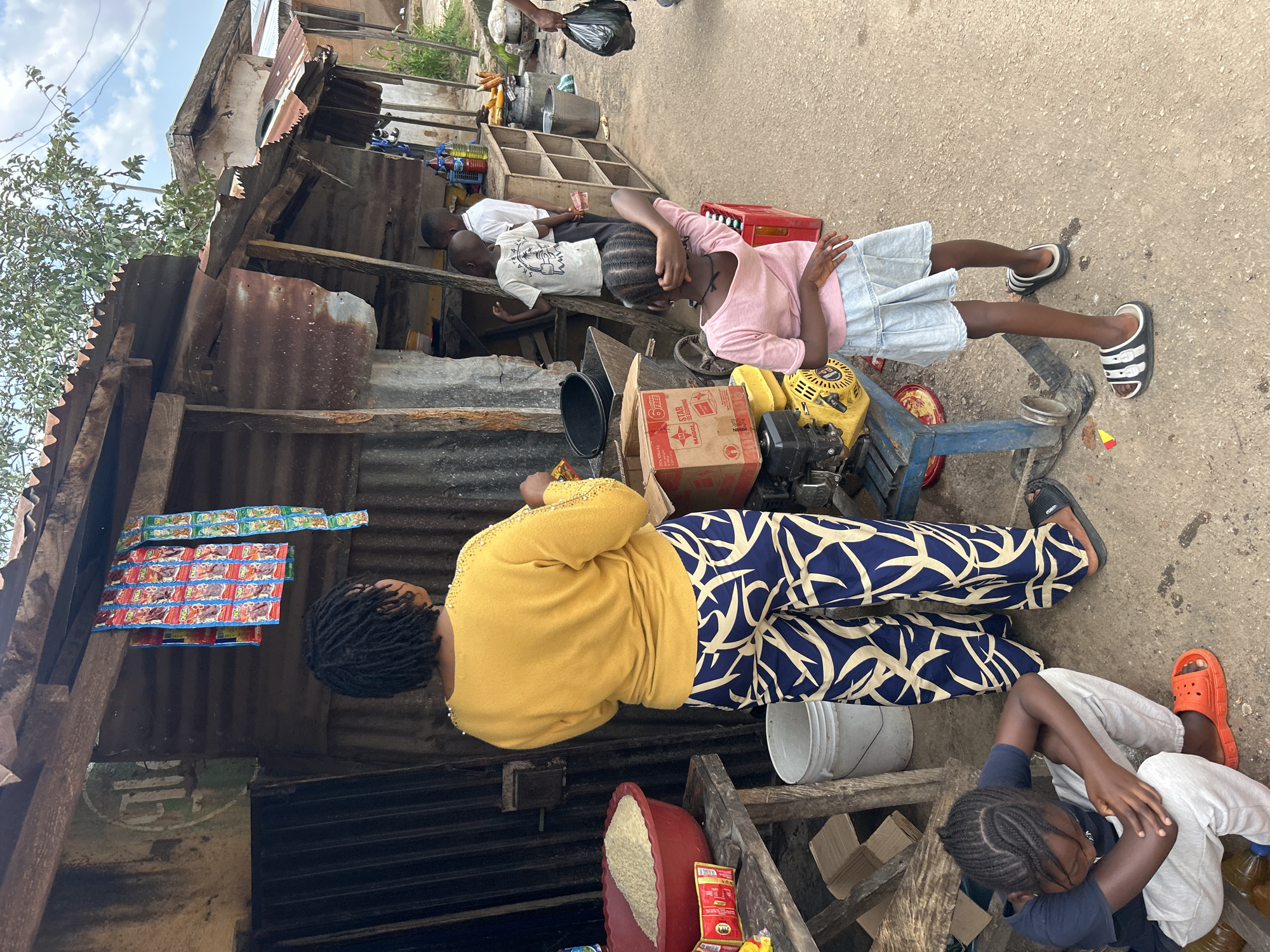 In the city of Jos, Nigeria, an outdoor market scene unfolds. A woman, wearing a yellow sweater and patterned pants, stands behind a table laden with goods. To her left, a large red basin overflows with rice, and several small packets of seasoning are scattered nearby. In front of her, a young girl in a pink t-shirt and a denim skirt stands, her back to the camera. To the right of the woman, a young boy in a white t-shirt and another person are partially visible. Behind them, shelves are stocked with various items, including bottles of liquid. A yellow generator is visible in the foreground. In the lower left corner, a child wearing a blue t-shirt and gray pants sits on the ground, looking down. The structures in the background are made of corrugated metal and wood, typical of makeshift market stalls.