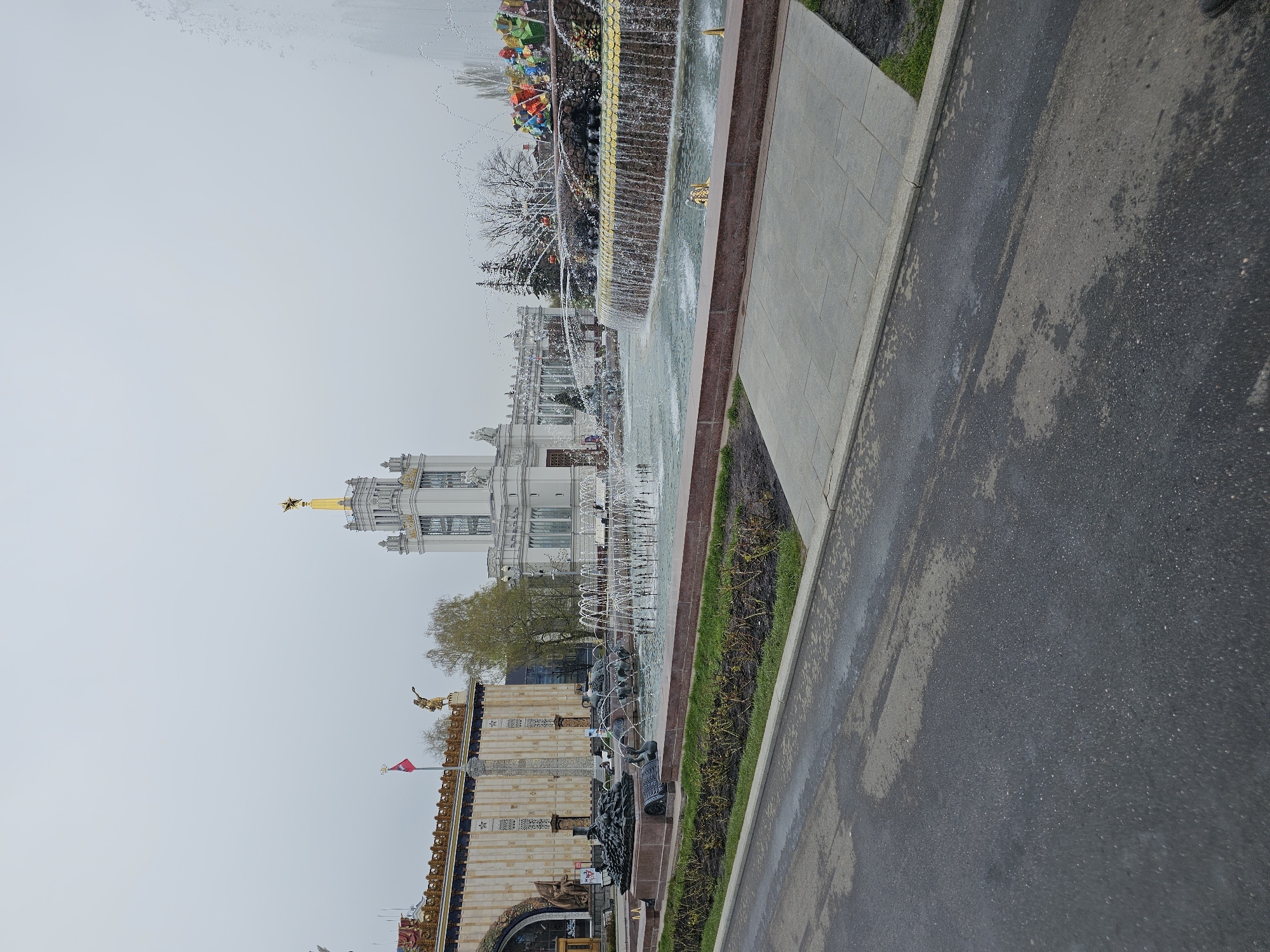 The image displays a vast outdoor scene within a grand architectural complex, characteristic of an exhibition center or park in Moscow. A wide, dark asphalt road or pathway, appearing damp in sections, runs along the right side of the frame, bordered by a concrete curb and a narrow strip of sparse green grass. Adjacent to the pathway, a large and ornate fountain complex dominates the mid-ground.
The multi-tiered fountain features numerous jets of water creating various patterns, with golden decorative elements and sculptures of figures, possibly representing allegorical or national symbols, placed around its basins. Further along the fountain, additional water features are visible, receding into the distance.
In the background, magnificent buildings with classical architectural styles rise under a uniformly grey, overcast sky. One prominent structure features a tall, golden spire crowned with a distinctive golden star. Another building closer to the foreground fountain is light-colored with elaborate detailing and has a small red flag flying from its rooftop. Scattered bare trees, some with initial leaf buds, suggest early spring or late autumn. The monumental scale of the architecture and fountains evokes a sense of historical grandeur. No individuals are visible, and no discernible text appears in the image. The weather appears damp and cool under the heavy cloud cover.