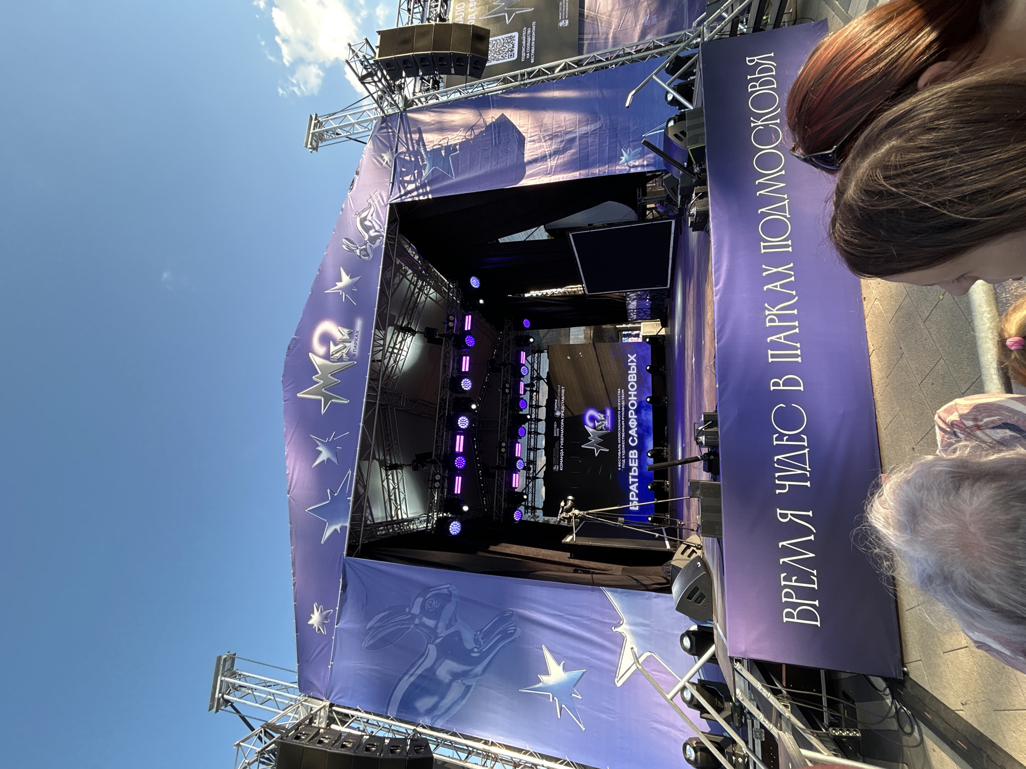 An outdoor stage is set up in front of a bright blue sky with scattered clouds. The stage is draped in purple fabric adorned with silver stars and a large silhouette of a rabbit. A large screen is visible behind the stage, displaying a logo and text. There are multiple speakers and lighting equipment on and around the stage. In the foreground, the backs of several people's heads are visible, suggesting they are part of an audience. The banner at the bottom of the stage reads "ВРЕМЯ ЧУДЕС В ПАРКАХ ПОДМОСКОВЬЯ" which translates to "Time of Miracles in Parks of Moscow Oblast". The context of the Moscow Oblast suggests this event may be taking place in a park within that region, potentially including Lyubertsy, Russia.