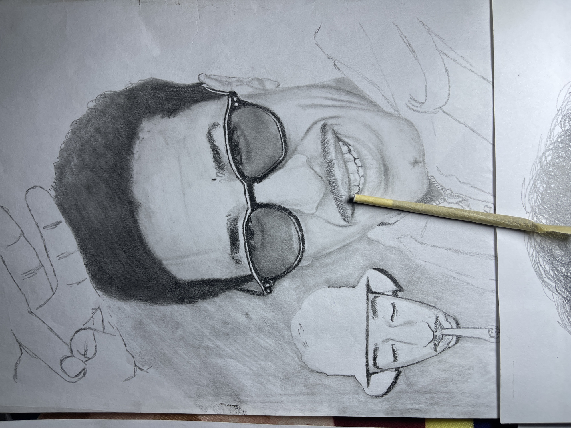 The image displays a detailed pencil drawing on a sheet of white paper, oriented vertically. The primary subject is a portrait of a smiling man with dark, wavy hair, wearing dark-framed sunglasses and a prominent mustache. His teeth are visible in the smile, and a light-colored chain or necklace is depicted around his neck. Above and to the left of the main portrait, a sketch of a hand with curled fingers is drawn, appearing to reach down. Below the main portrait's left shoulder, a smaller, less detailed drawing of a man wearing a bucket hat, with closed eyes and a goatee, is depicted. This smaller figure holds a thin, tapered object between his lips, resembling a pipe or cigarette. A light-colored, slender cylindrical object, possibly a pencil or a rolled paper blending stick, extends diagonally from the lower left towards the upper right, partially obscuring the main figure's mouth and mustache. The drawing paper shows minor creases and smudges, consistent with active use. The top drawing partially overlays another sheet of white paper at the bottom, revealing an additional, unfinished sketch of hair or scribbles. A faint multi-colored stripe (red, yellow, blue) is visible along the left edge beneath the primary drawing, suggesting the surface on which the artwork rests. The visual content of the image solely depicts artwork and does not contain any discernible environmental features or landmarks that would identify a specific physical location within Jos, Nigeria, or any other city.