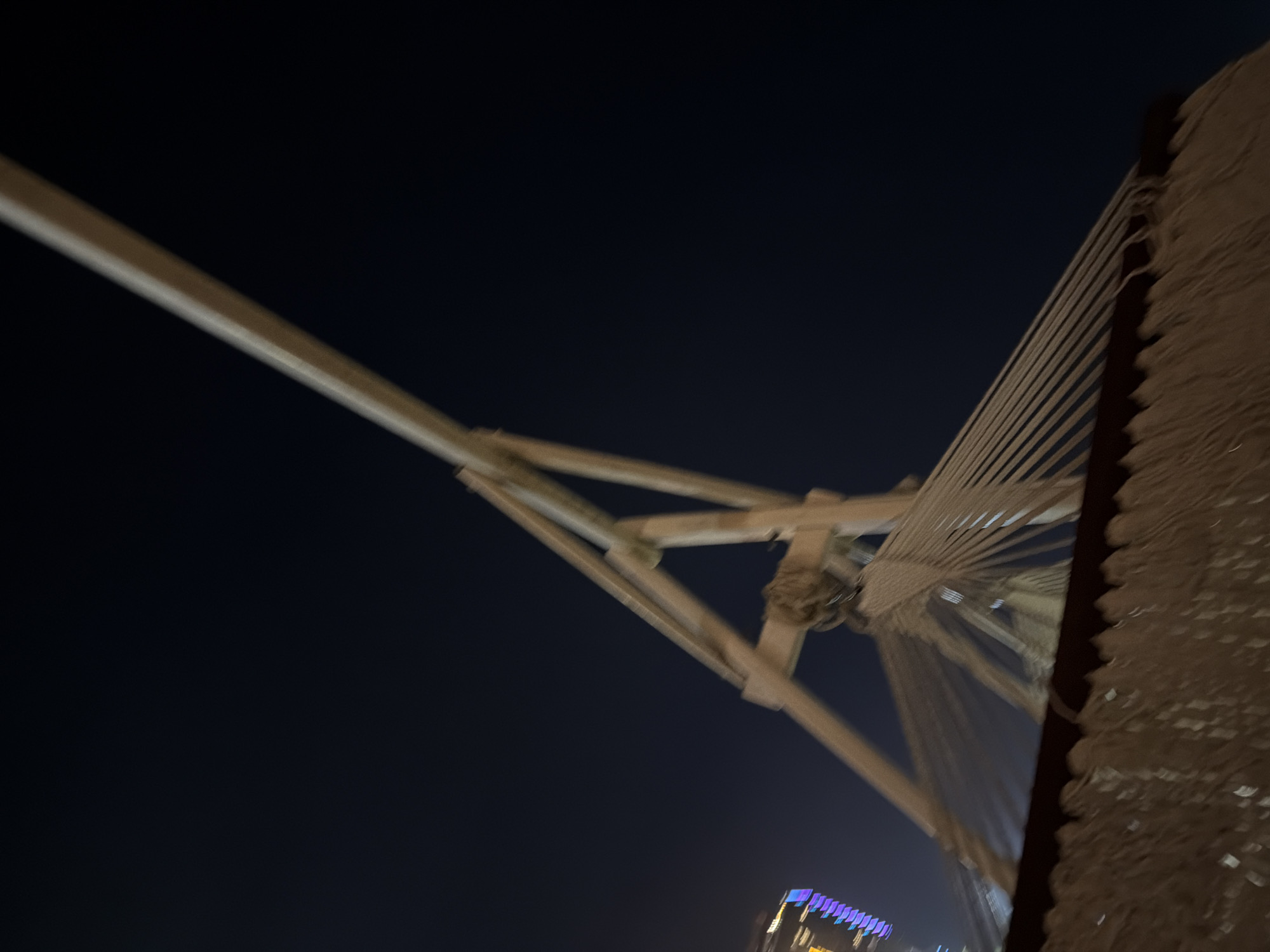 The image captures a striking architectural structure against a dark night sky, likely a large bridge, viewed from below. A prominent, light-colored diagonal beam extends from the upper left towards the center, where it connects with other beams, forming a truss-like support system. From this central connection point, numerous thin, parallel cables fan out, descending towards a textured, vertical support on the right side of the frame. This vertical element appears rugged and textured, possibly covered in concrete or stone.
The overall scene is illuminated, though the light source is not visible, suggesting artificial lighting on the structure itself. The sky is a deep, dark blue or black, indicating nighttime. In the bottom right corner of the main image, distant city lights are visible, illuminating a building with distinct purple and blue hues, hinting at a bustling urban environment.
There are no people visible, and no specific activity is taking place; the image is a static architectural shot. The structure's design, particularly the radiating cables and angular framework, strongly suggests a cable-stayed bridge. Given the location context of Đà Nẵng, Vietnam, it is highly probable that this is a detail of the iconic Trần Thị Lý Bridge, known for its distinctive sail-like mast and cable design. No visible text is present in the image.