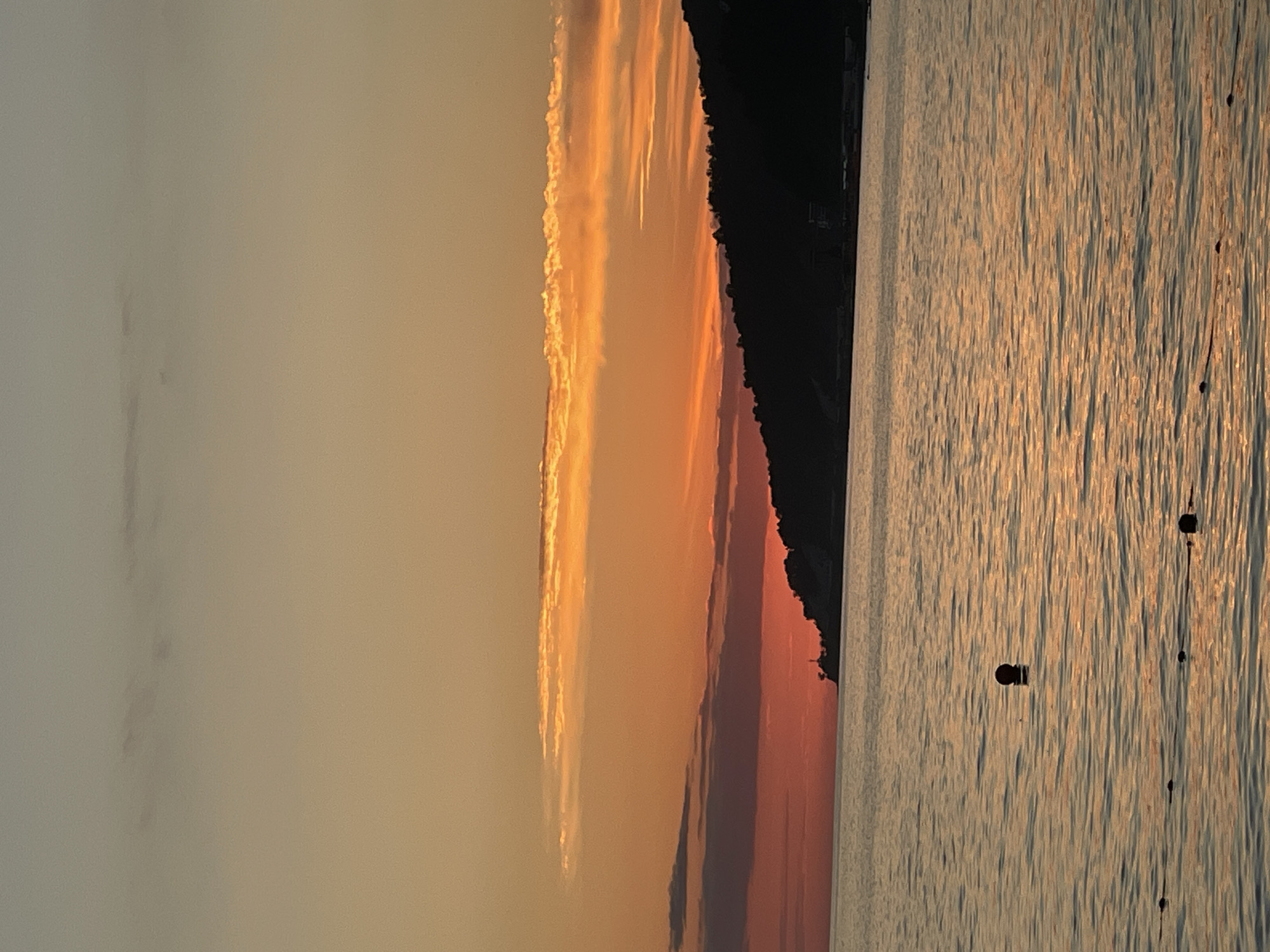 A serene sunset is captured over the Black Sea, with the silhouette of a hilly coastline visible on the right. The sky transitions from a soft grey at the top to vibrant orange and red hues closer to the horizon. Wispy clouds are scattered across the sky, catching the warm light of the setting sun. The sea is calm, reflecting the colors of the sky with gentle ripples. A few dark buoys or markers are visible floating on the water's surface, suggesting a protected swimming area or a navigational marker. In the distance, on the coastline, a small lighthouse or tower-like structure is faintly discernible. The overall scene evokes a peaceful and tranquil atmosphere. The scene is likely located along the coast of Sochi, Russia, given the characteristic hilly terrain meeting the sea and the typical cloud formations and lighting conditions often seen at sunset in this region.