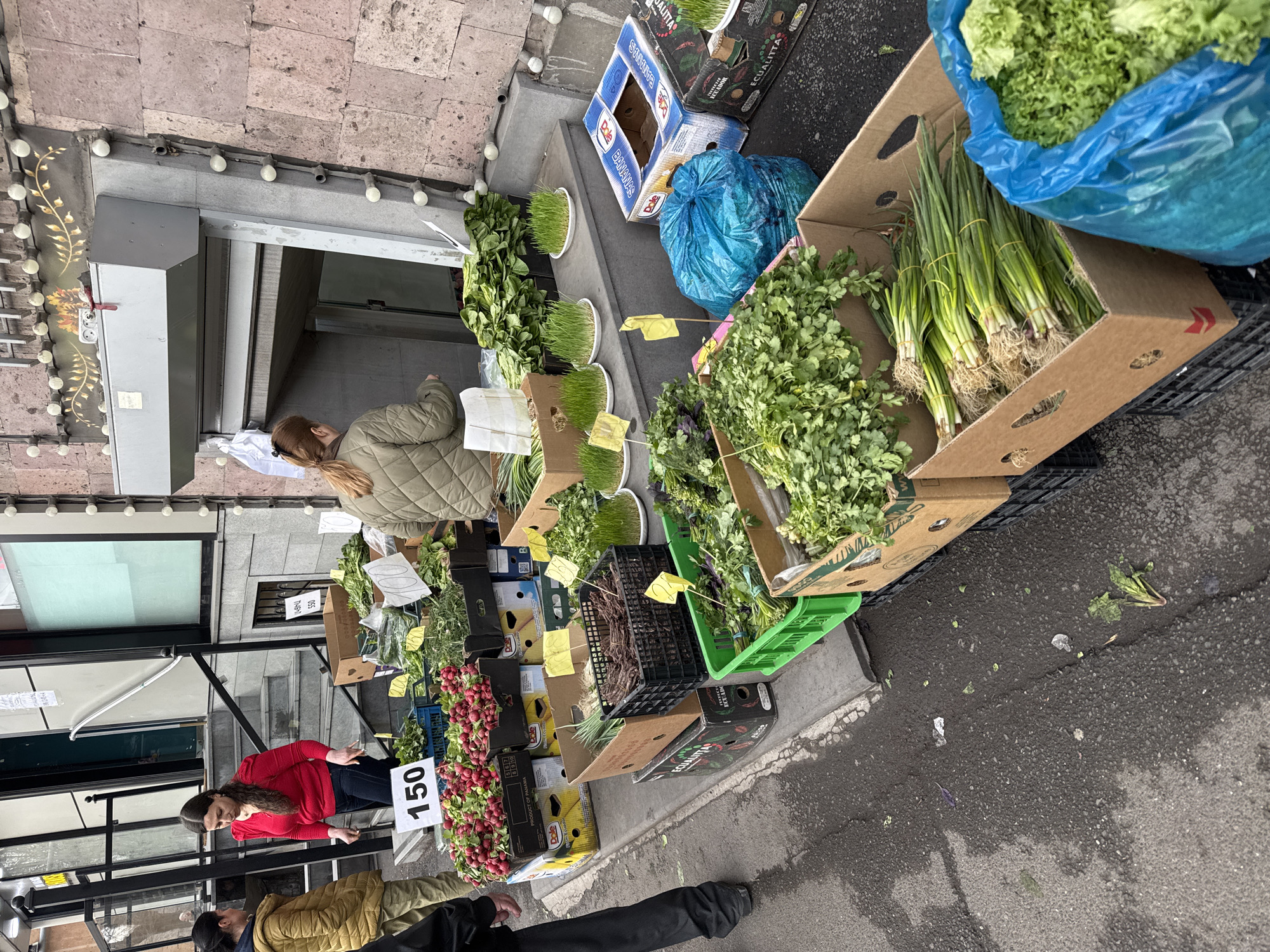 This image captures an outdoor market scene, likely in Yerevan, Armenia, displaying various fresh produce items for sale. The scene is shot from a slightly elevated perspective, oriented horizontally, but with a counter-clockwise rotation. The foreground features radishes with a sign indicating a price of 150 (likely Armenian dram). A vendor in a red top is positioned near the radishes. Nearby, a variety of other greens and vegetables are displayed in boxes and crates, including what appear to be cilantro, green onions, and lettuce. The boxes and crates are stacked on the sidewalk. In the background, additional produce is visible, along with customers browsing the market. The backdrop includes buildings with windows and architectural details. The sidewalk surface appears wet.