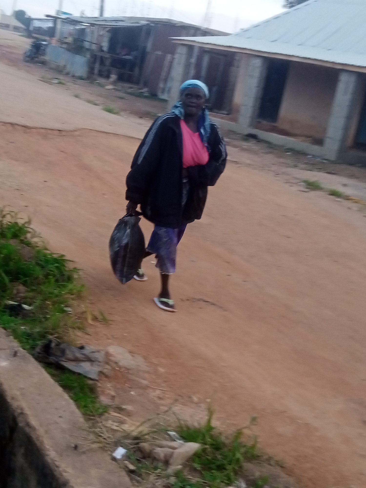 A woman is walking on a dirt road in Russo, Nigeria. She is wearing a blue headscarf, a pink t-shirt, and a dark jacket. She is carrying a large black bag in her left hand and wearing sandals on her feet. The road appears to be unpaved and dusty. In the background, there are some buildings that appear to be made of corrugated metal and concrete. There is also some vegetation along the side of the road. The sky is overcast, suggesting it might be early morning or late afternoon, or a cloudy day. The overall impression is a simple, rural scene.