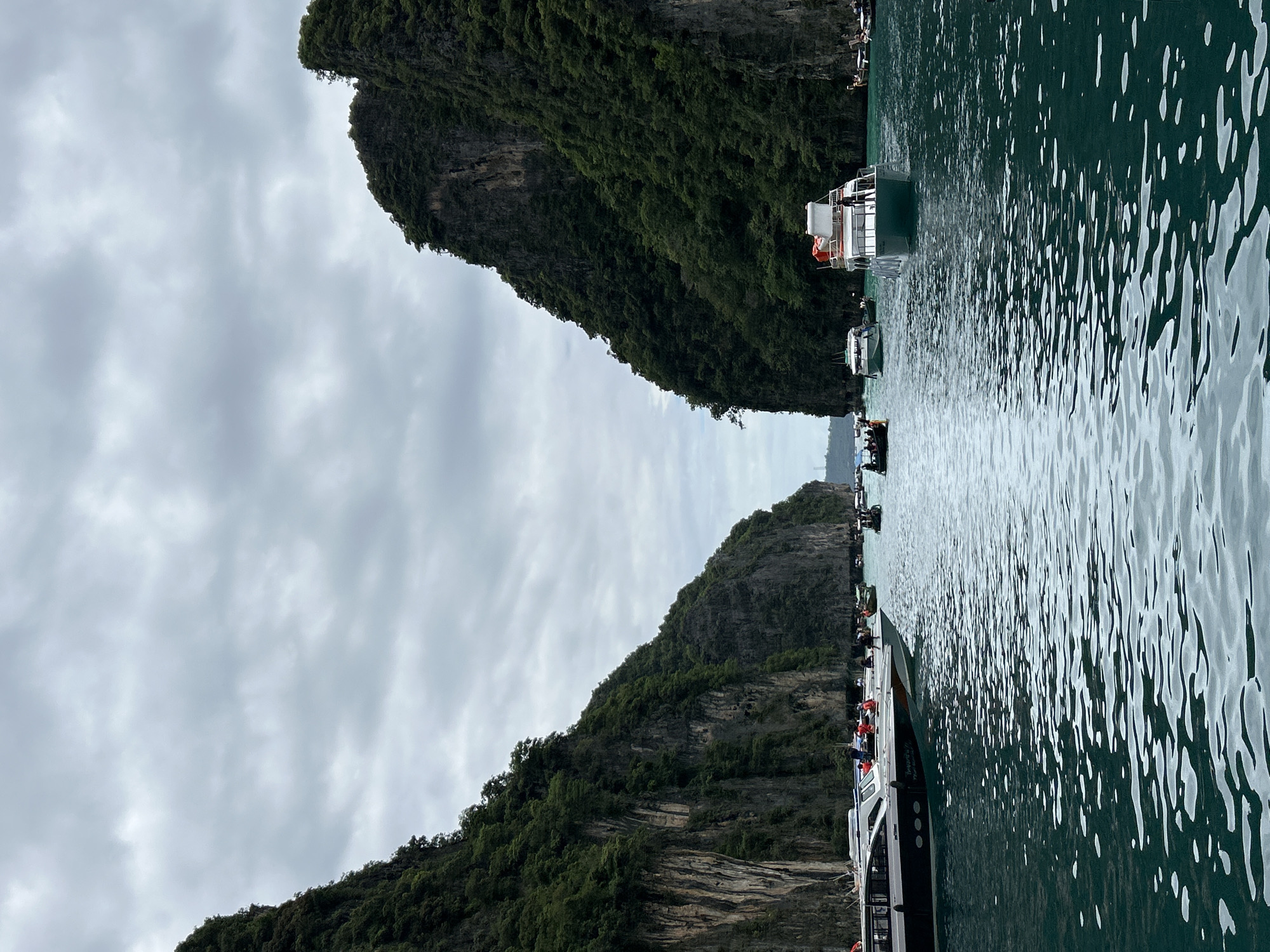 The image depicts a view of the turquoise waters of Ao Nang, Thailand, as seen from a boat. Towering limestone cliffs covered in lush green vegetation flank a narrow strait, creating a dramatic natural passage. Several boats of various sizes are present, including speedboats and smaller vessels, with people visible on their decks, suggesting a popular tourist destination. The water exhibits a rippling texture, reflecting the overcast sky filled with grey clouds. The scene captures a moment of serene yet active marine traffic amidst a breathtaking natural landscape.