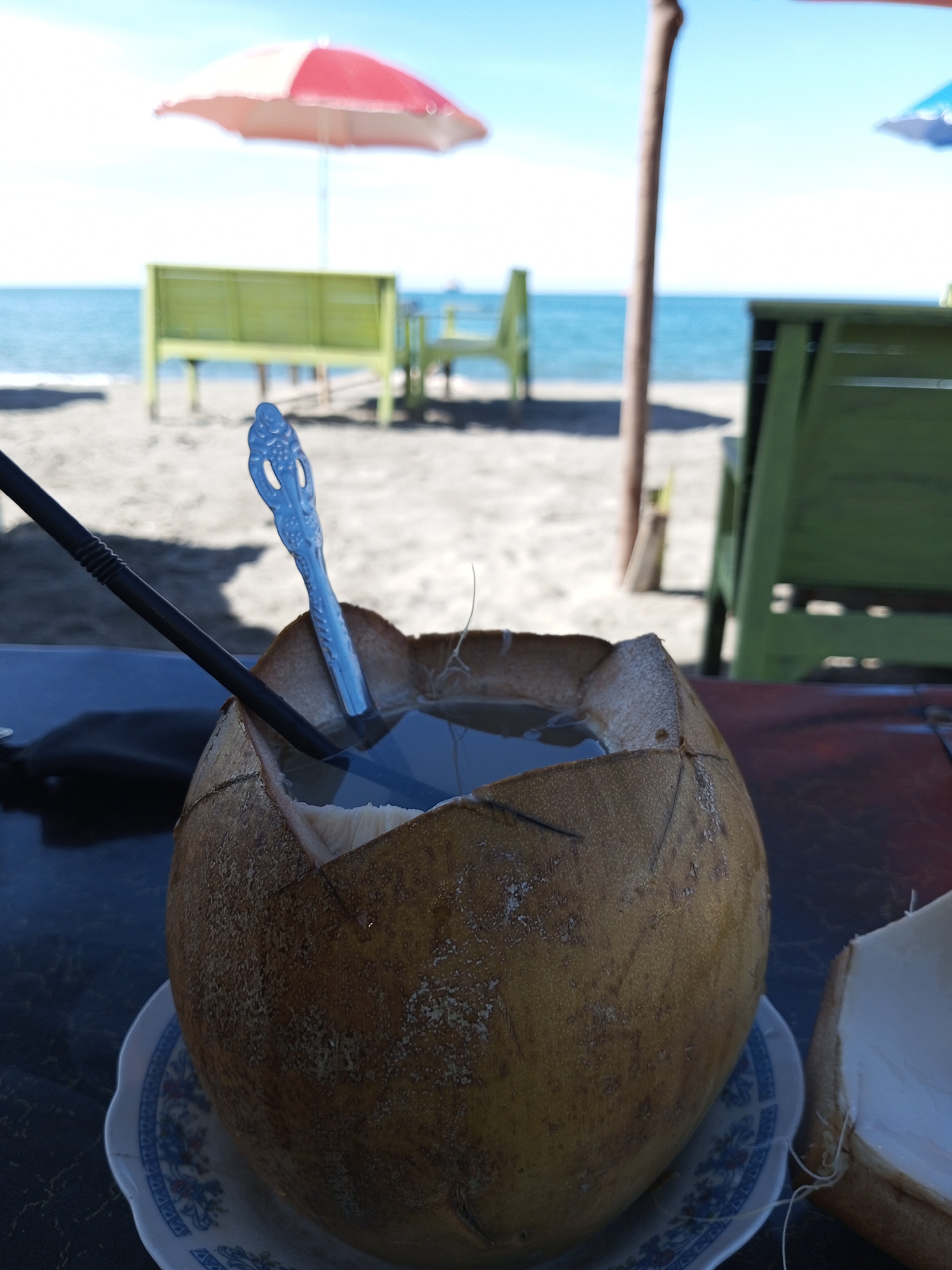 The image captures a refreshing coconut drink on a beach in Lhokseumawe, Indonesia. The foreground features a halved coconut filled with water, placed on a decorative plate. A black straw and a blue plastic spoon are inserted into the coconut. Behind the coconut, green wooden benches and tables are visible on the sandy beach. In the background, there is a view of the sea, a beach umbrella with red and white panels, and a clear, bright sky.