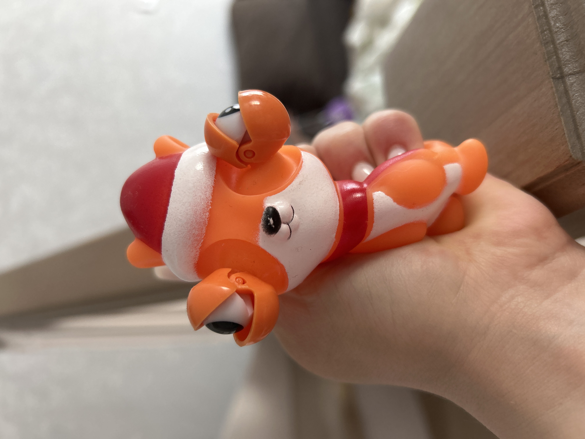 A hand holds a small, orange and white plastic toy resembling a clown. The toy features articulated eyeballs within orange casings that can open and close. Red stripes are painted around the toy's head and midsection. The background includes a blurred wooden surface and a neutral-toned wall. The location in Chaikovsky, Russia is not discernible from the image.